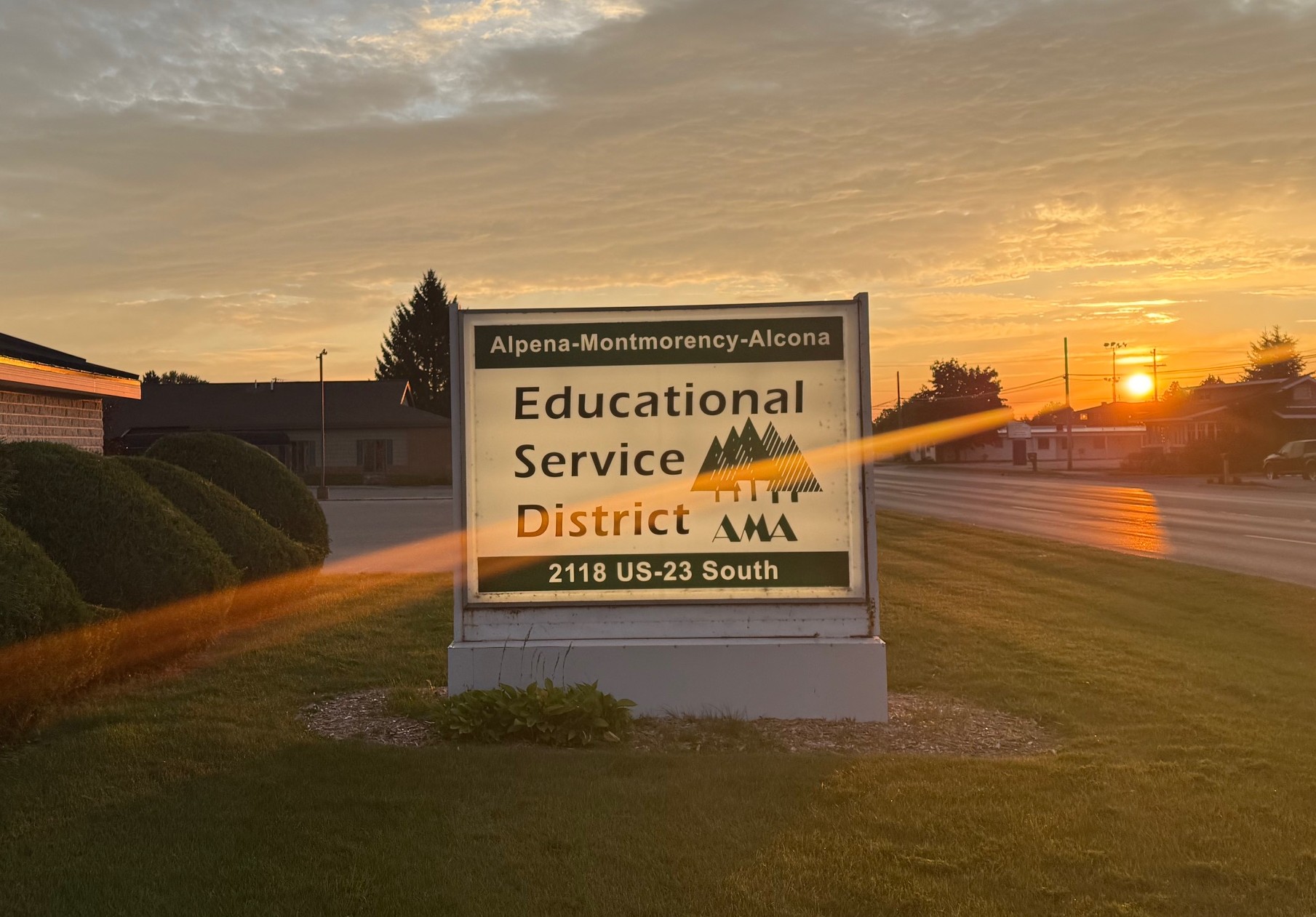  large sign for the Alpena-Montmorency-Alcona Educational Service District, or AMA ESD, stands in a grassy area at sunrise. The sun is visible just above the tree line in the background, casting an orange glow and a sunburst effect across the sky and the sign. The sign itself is lit from within, with the words "Alpena-Montmorency-Alcona" at the top and "Educational Service District" written in larger letters below. The AMA logo, which features a tree and mountain design, is also visible on the sign. The address, 2118 US-23 South, is at the bottom of the sign.