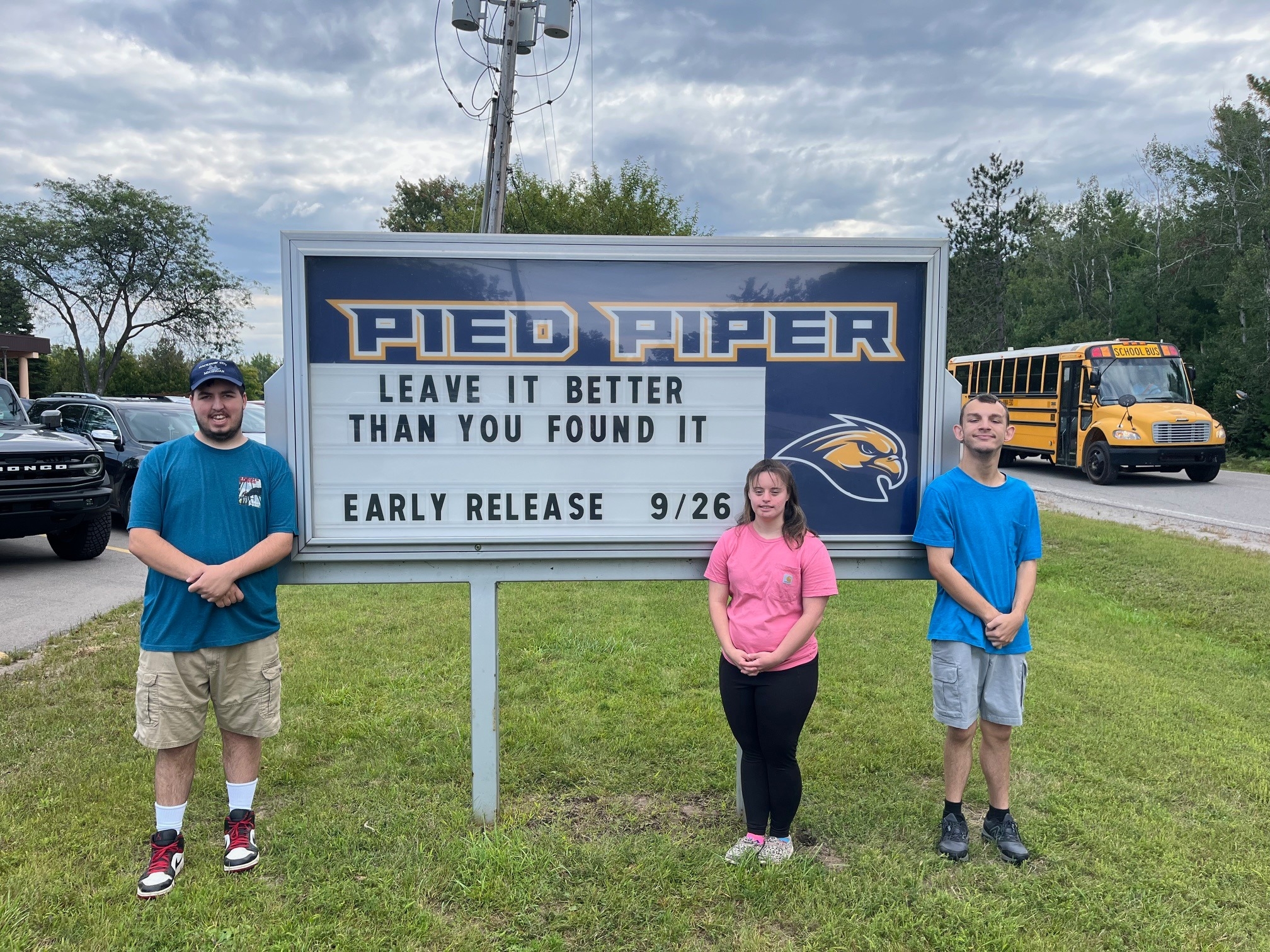 A wide-angle, full-body outdoor shot shows three young people, a man on the left, a woman in the center, and a man on the right, standing side-by-side in front of a large sign for Pied Piper, which features a golden eagle logo. The sign has a message board that says "Leave it better than you found it" and "Early release 9/26". A Ford Bronco is partially visible on the far left, and a school bus is on the far right. The people are smiling slightly and looking at the camera. The sky is partly cloudy, and the grass is green.