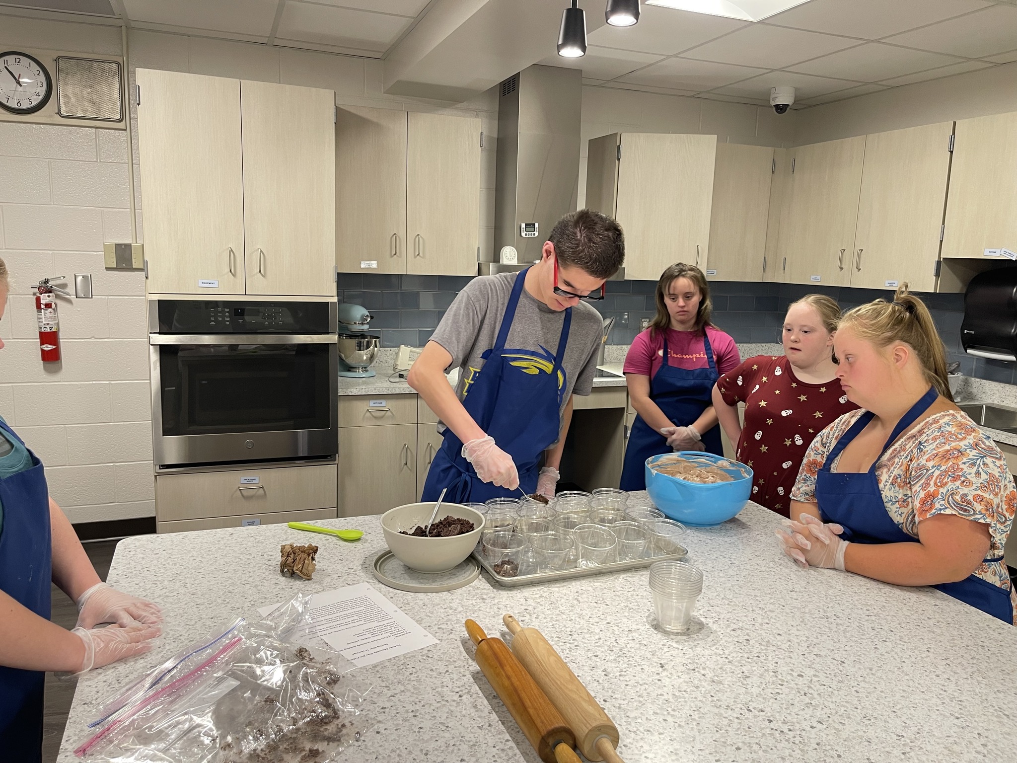 A group of students wearing blue aprons and gloves are working together in a school kitchen. One student in the center is carefully scooping ingredients into clear cups while others watch and assist. Bowls of ingredients, rolling pins, and a recipe sheet are spread across the counter. Light wood cabinets and a stainless-steel oven are in the background.