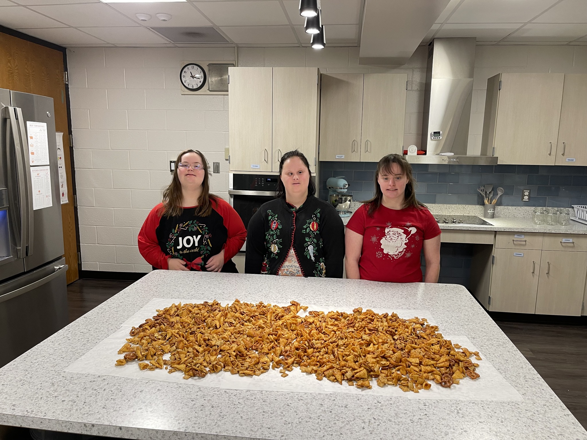 Three young women wearing holiday-themed shirts stand behind a large pile of golden-brown dried fruit, likely apples or peaches, on a kitchen counter.