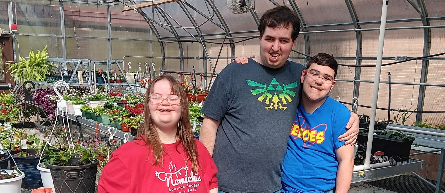 Three young adults—two men and one woman—smile together inside a sunlit greenhouse. The woman on the left wears a red t-shirt and patterned leggings, holding a green watering can. The man in the center wears a dark grey t-shirt with a green graphic, and the man on the right wears a bright blue t-shirt; they stand with their arms around each other. Rows of potted plants and flowers on metal benches fill the background under the arched greenhouse structure.