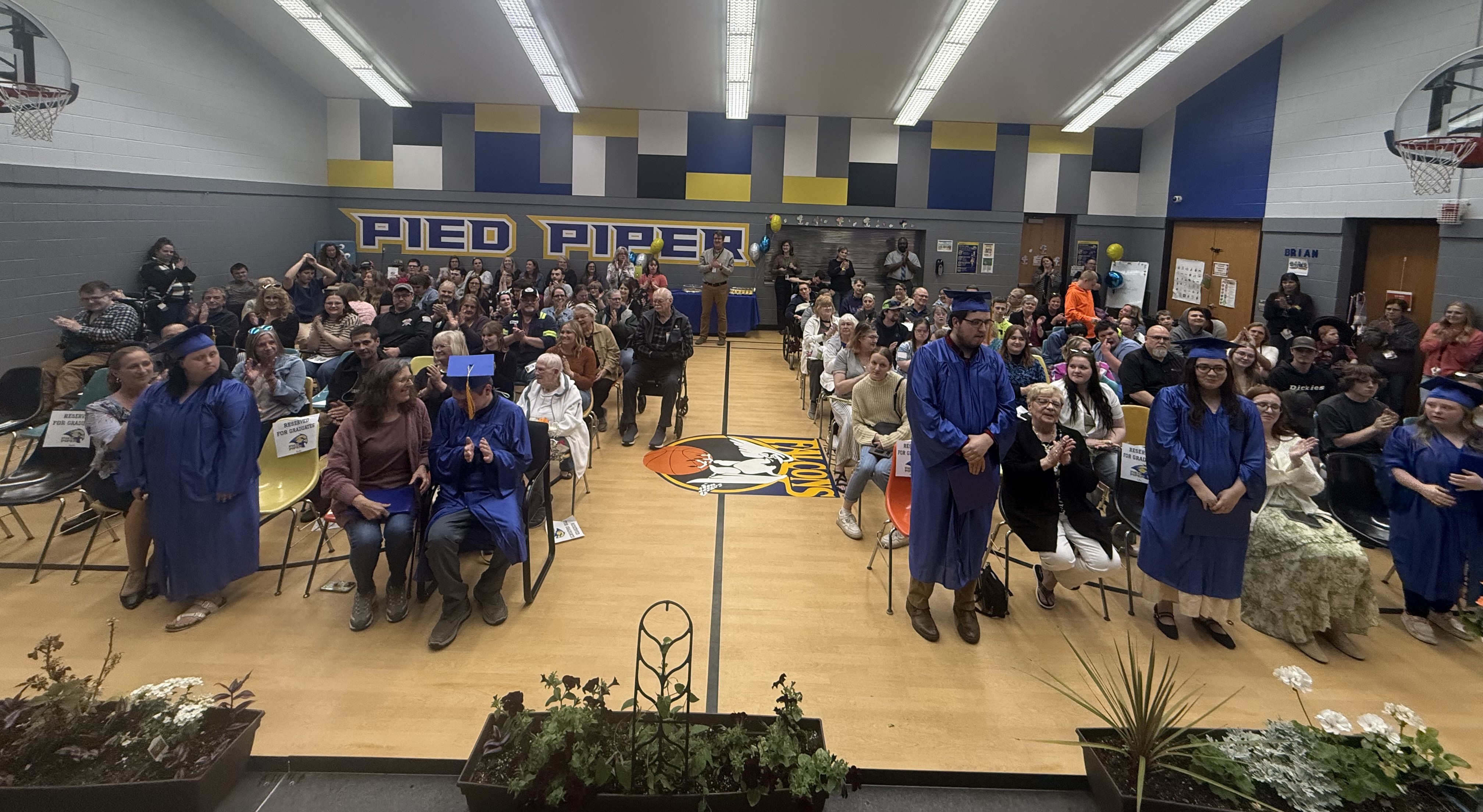 Graduates in blue caps and gowns stand proudly in a decorated school gymnasium during a Pied Piper graduation ceremony, surrounded by rows of applauding family members, friends, staff, and community supporters. The room features blue, gold, gray, and white school colors, basketball hoops, and a large “Pied Piper” mural on the back wall, capturing a warm and celebratory atmosphere.