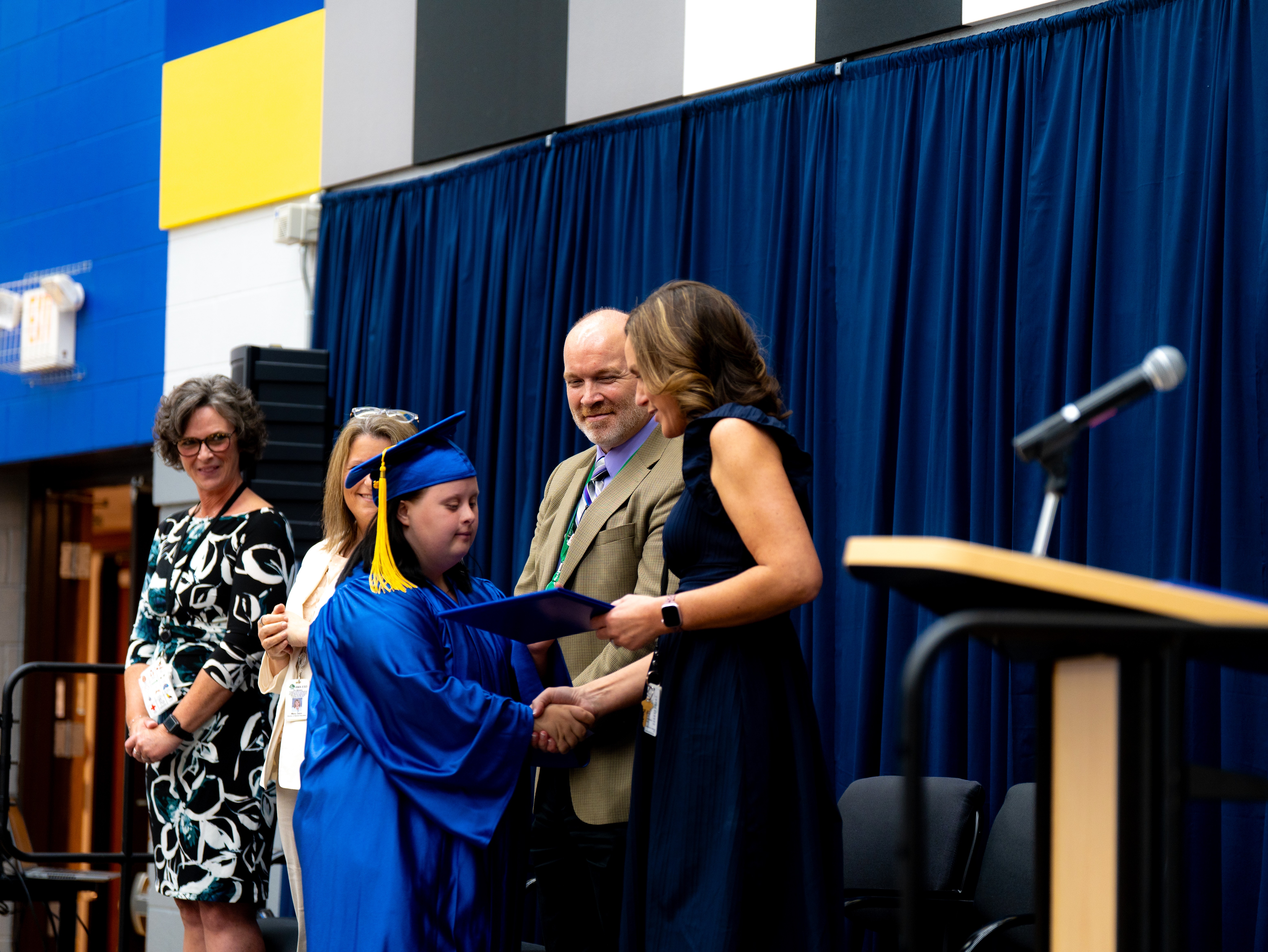A graduate in a blue cap and gown shakes hands with a staff member while receiving a diploma folder on stage during a graduation ceremony, with other staff members, a podium, microphone, and blue curtains in the background.