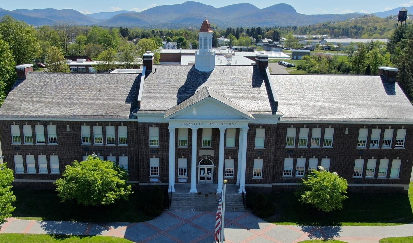 This is a picture of front of Granville Jr/Sr High Building - a building with four pillars, a flag out front and the Vermont Green Mountains in the background