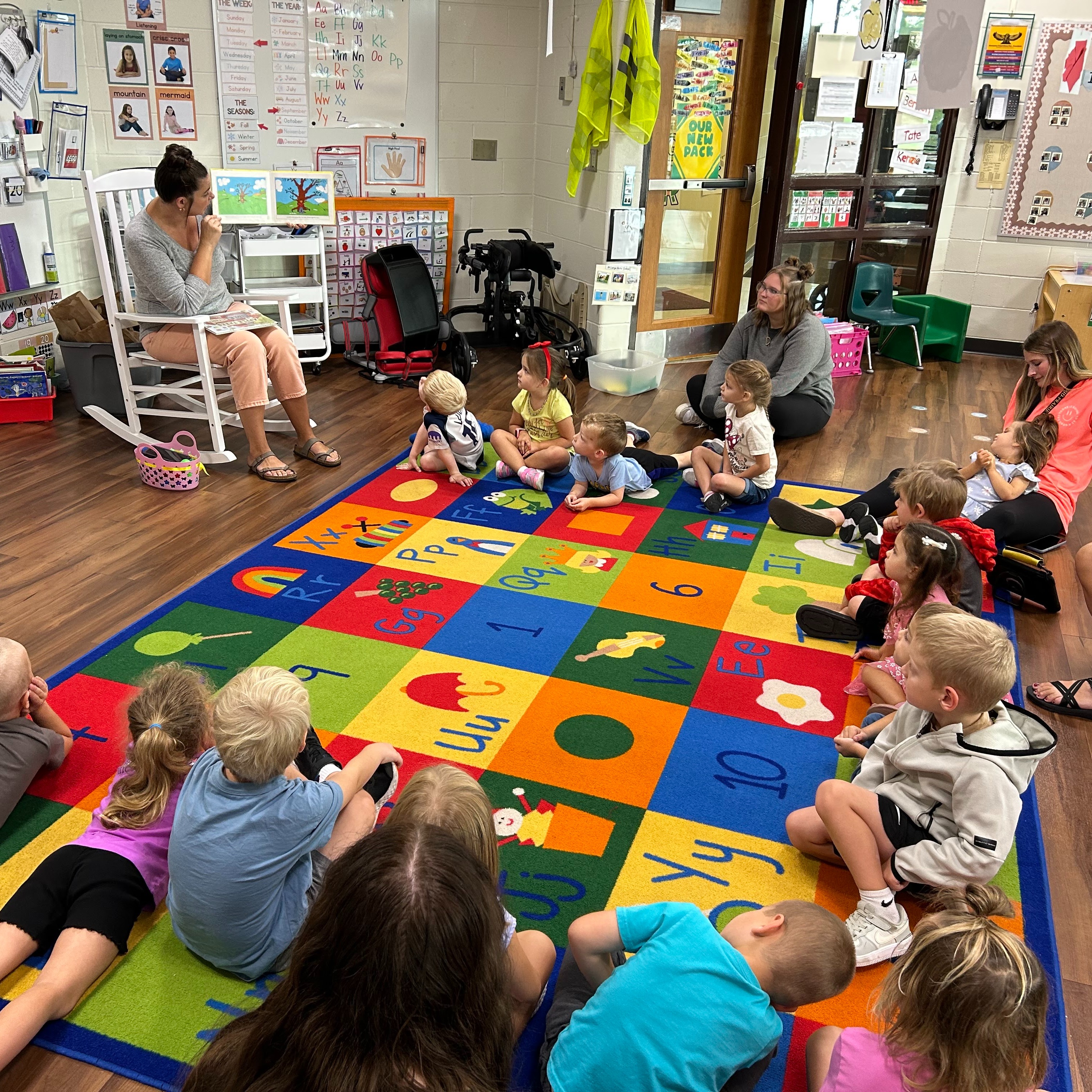 preschol Students in class listening to an adult reading a story aloud at the front of the room; carpet text reads the A B Cs