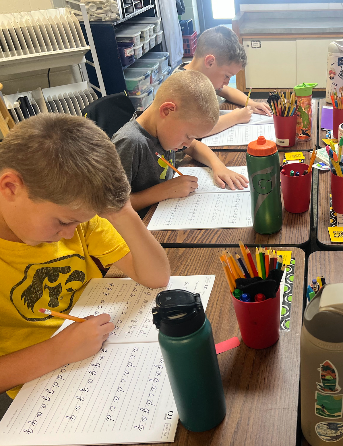 Students sitting at desks with pencils and a cursive practice sheet writing the letters a b c d e f g repeatedly