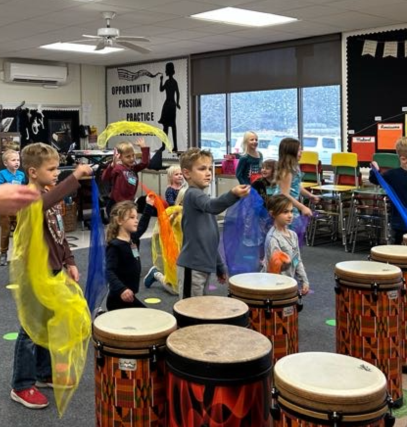 Students standing in the classroom with colorful scarves throwing them up into the air with drums in front of them in the classroom