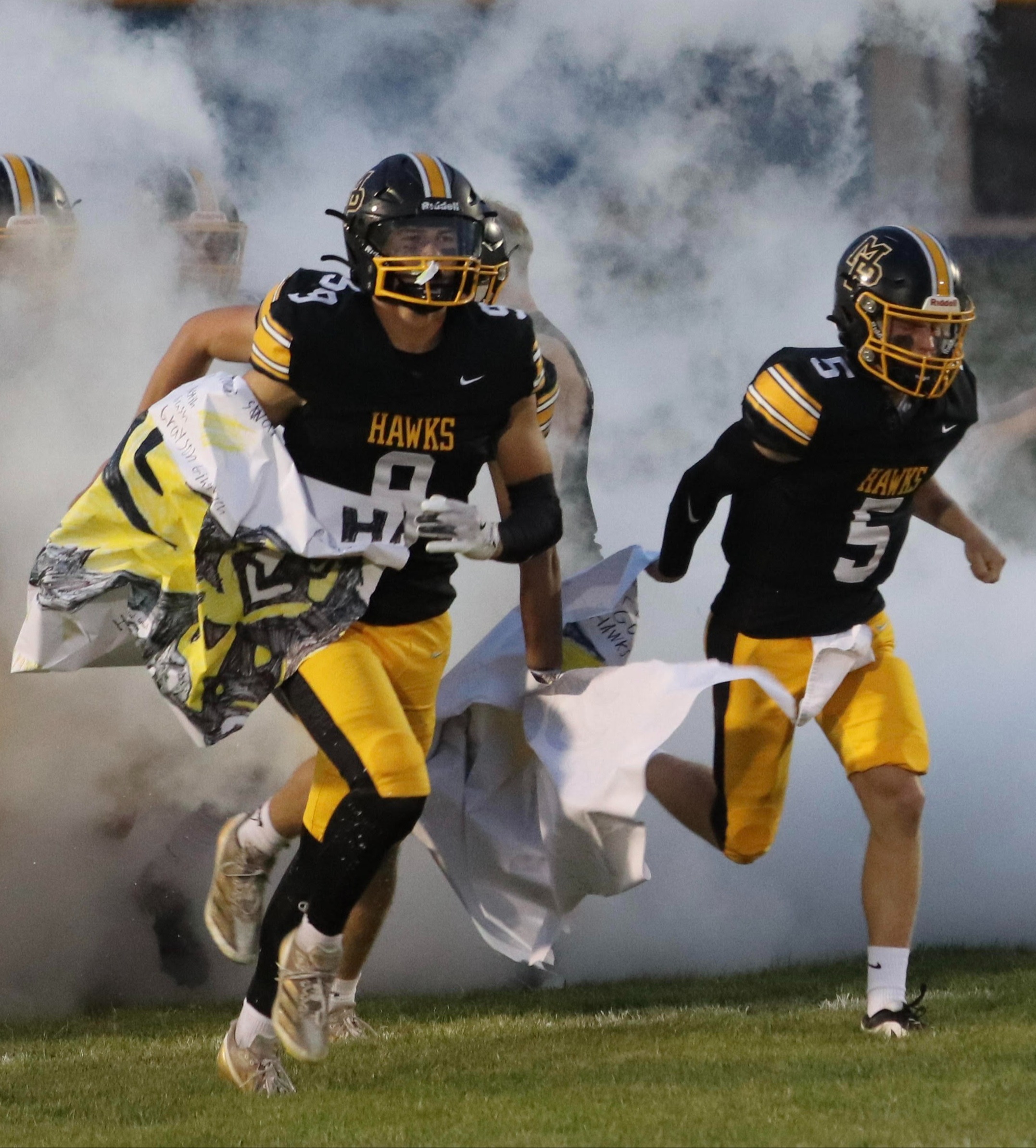 Two football players in black and gold uniforms with Hawks written on the uniforms and numbers 9 and 5 running out onto the field through smoke