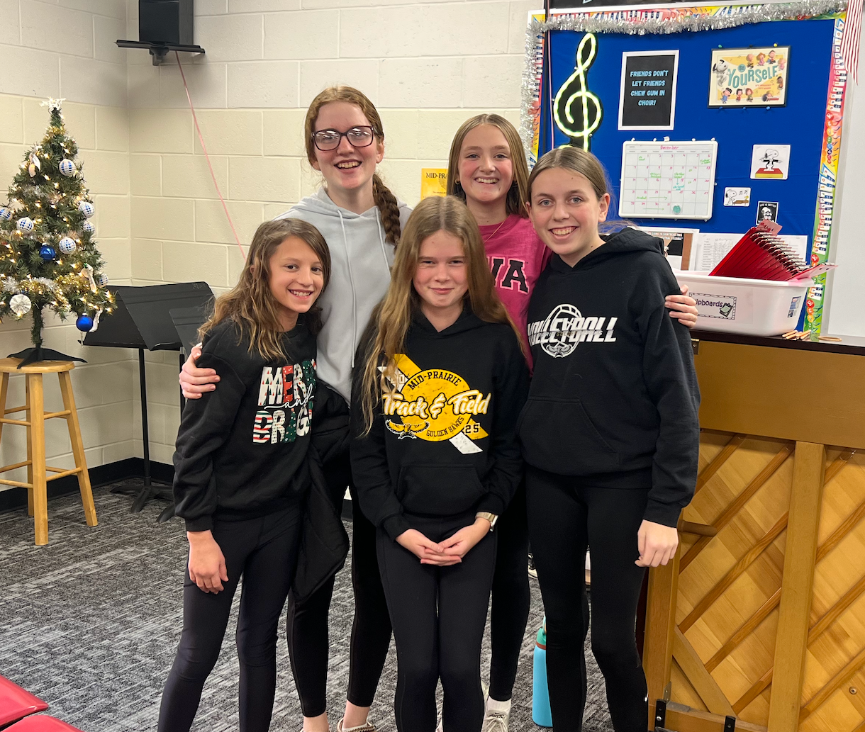 Five female students standing in the music room in front of the piano smiling with their arms around each other; Wall behind reads Be Amazing Today