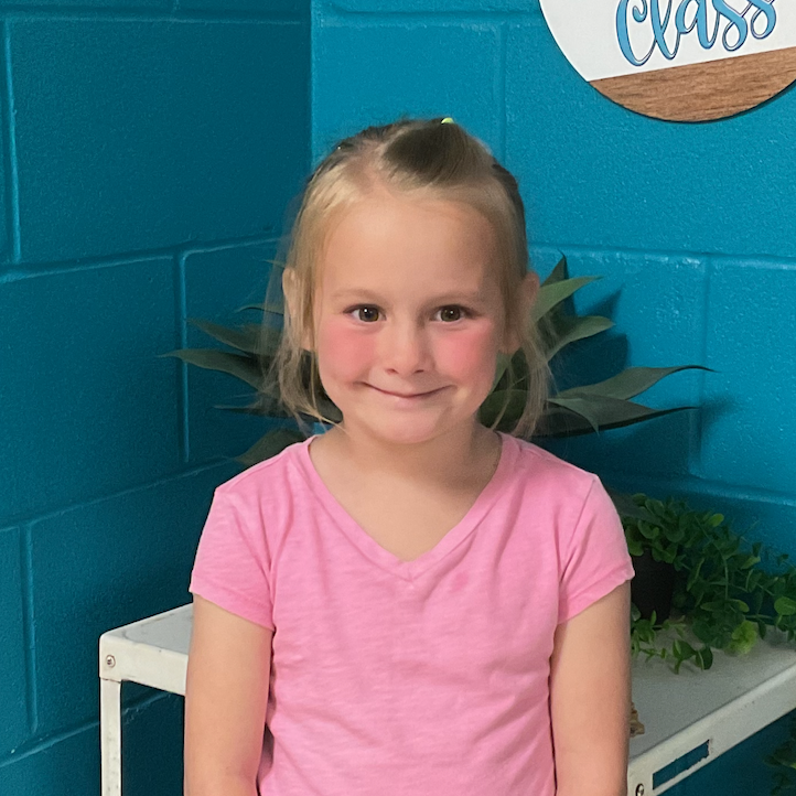 Molly Yoder wearing a pink t shirt standing in front of a turquoise wall and plant and a sign that says Welcome to our class