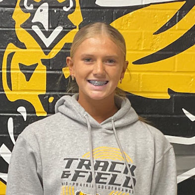 Tessa wearing a grey sweatshirt that says Track and Field standing in front of the black and gold MP hawk wall decal