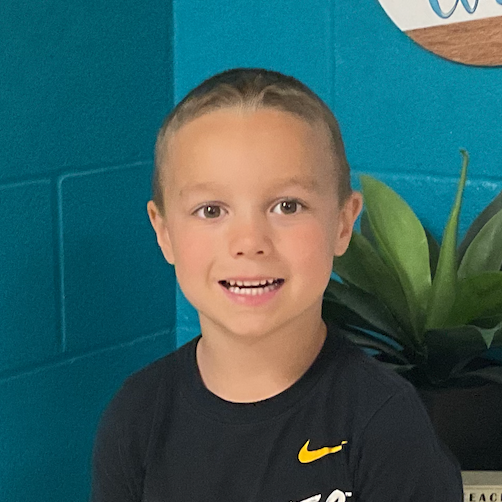 Miles Berg wearing a t-shirt that says HAWKEYES standing in front of a Welcome to our Class sign and a block that reads Teachers who love teaching, teach children to love learning