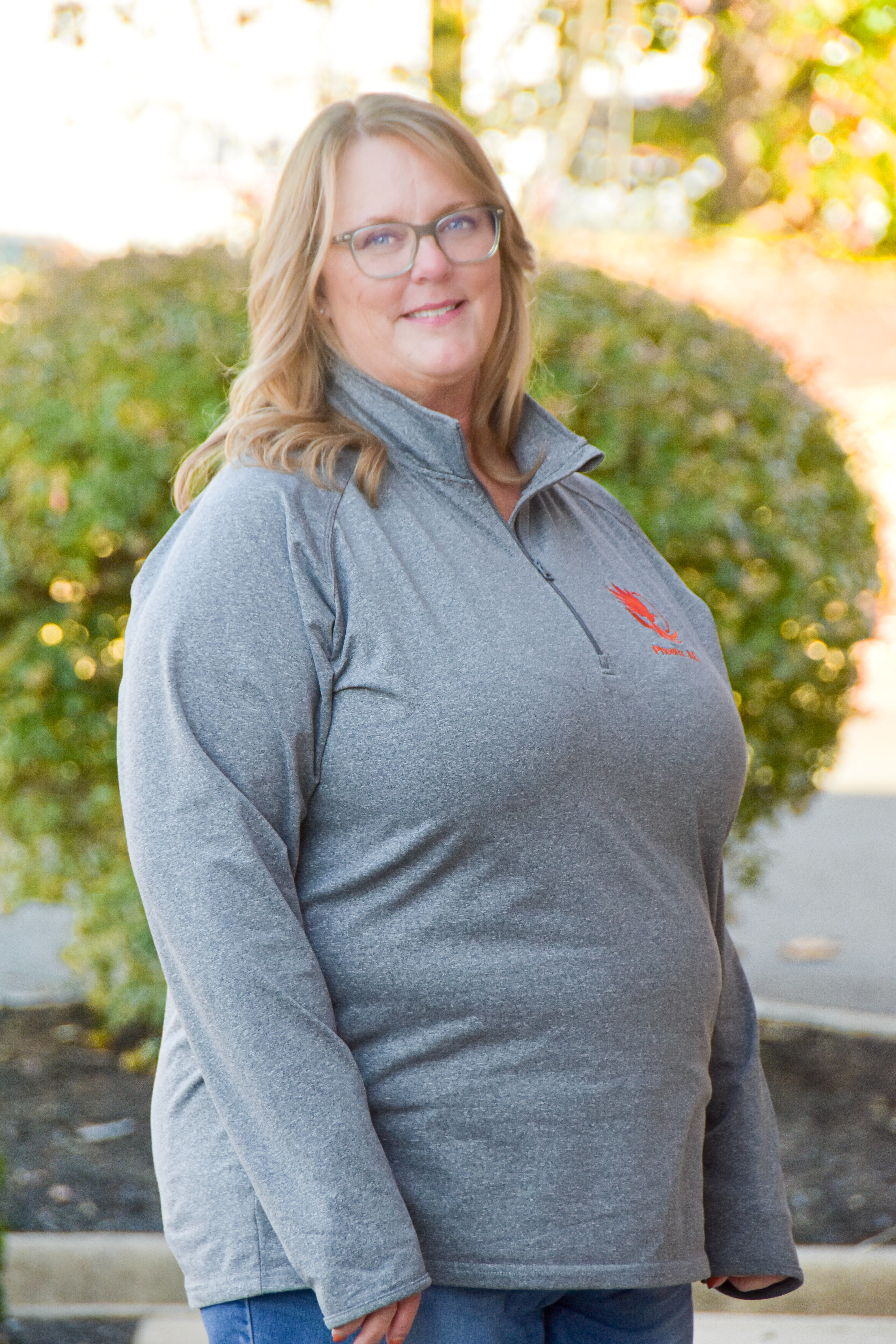Woman with blonde hair wearing glasses, a gray shirt, and blue jeans. Behind her is greenery.