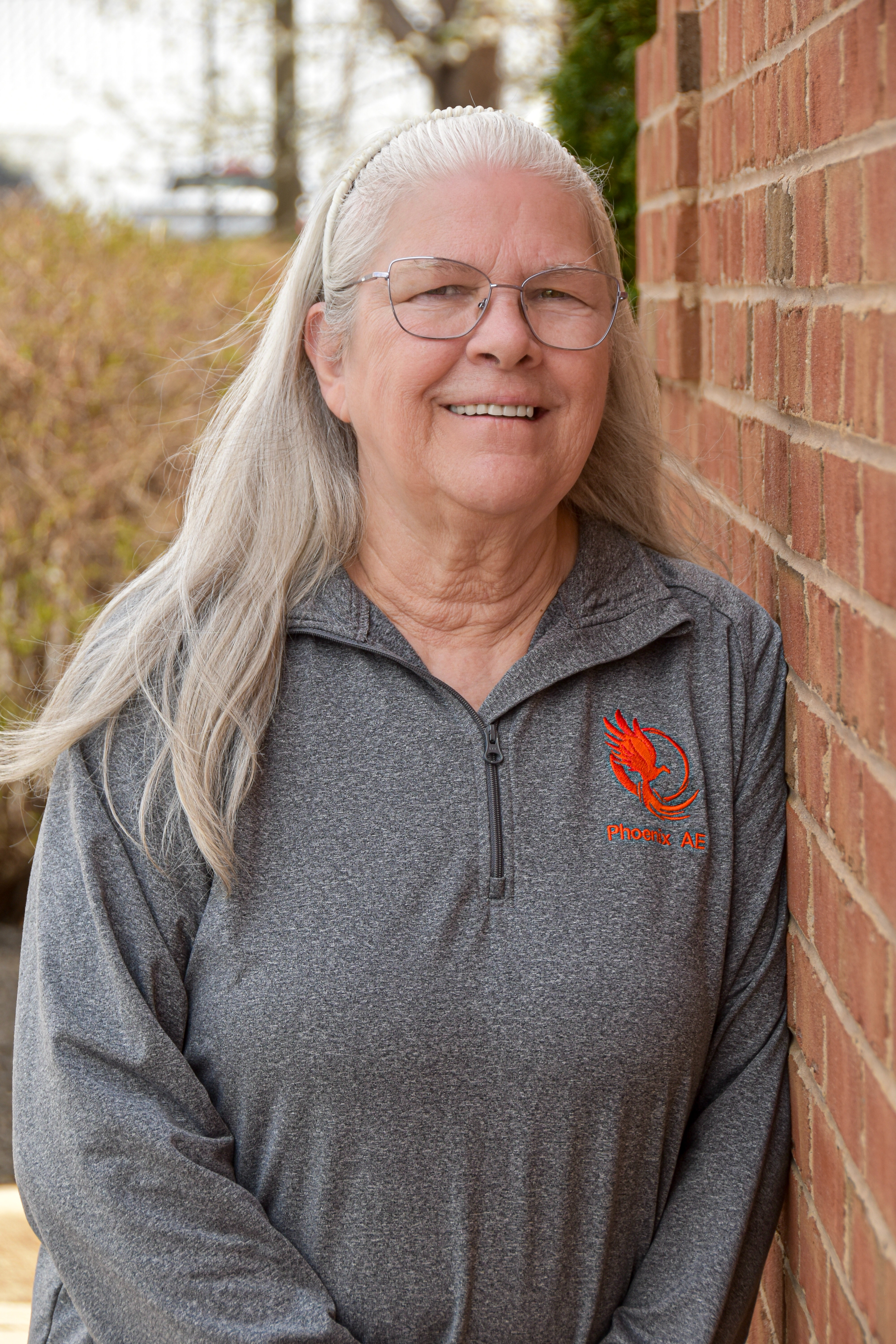 Woman with long gray hair in a gray long-sleeved shirt. She wears glasses and stands by a brick wall.