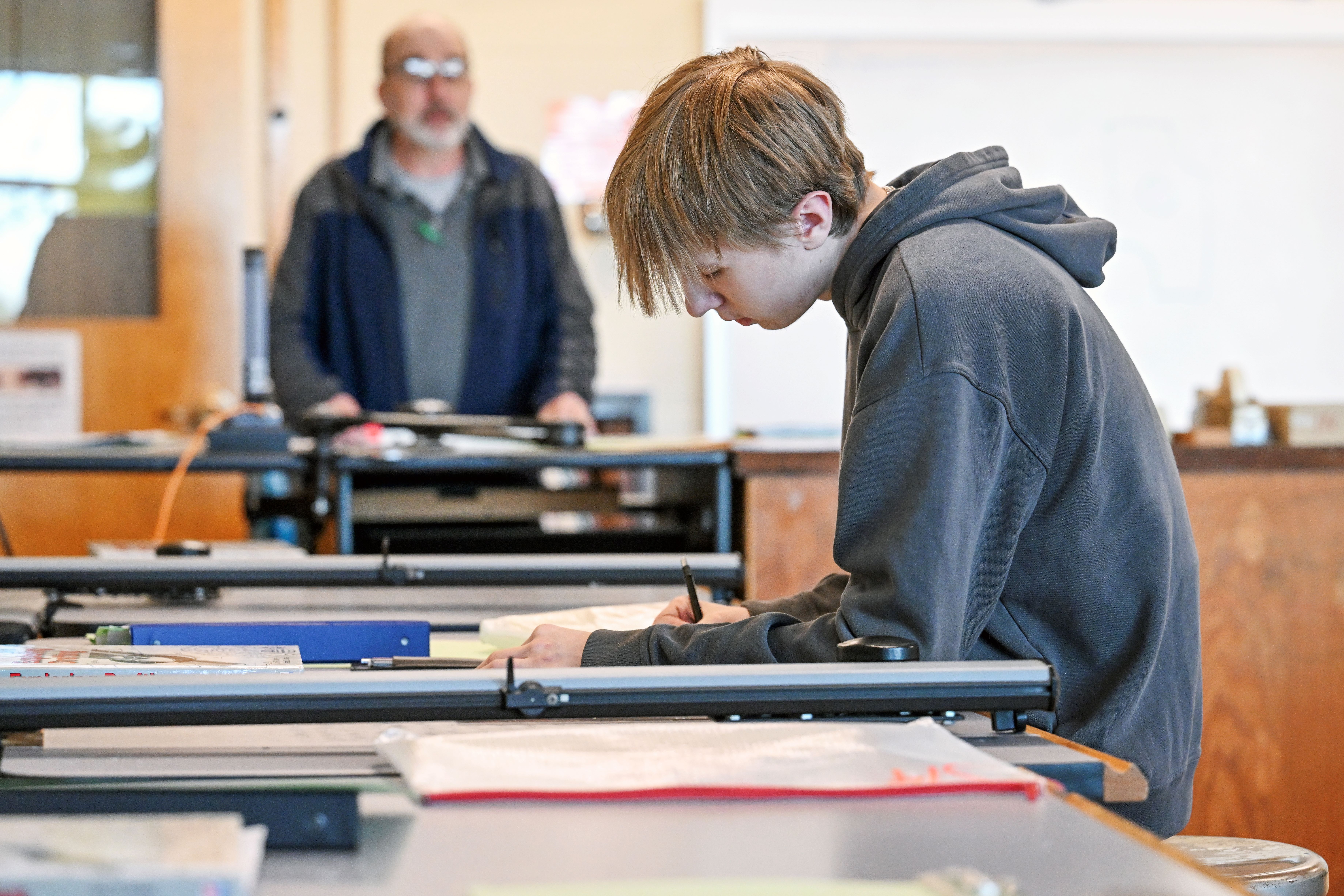 Male student working on a project with a male teacher looking on in the background.