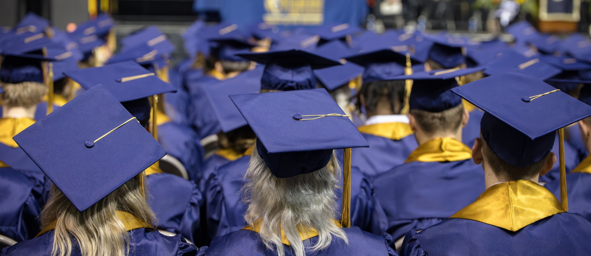 Palmer Ridge High School Class of  2024 dressed in blue graduation caps and gowns with gold accents sit in rows facing a stage during a graduation ceremony. The stage in the background has speakers, decorations, and a table with a banner.