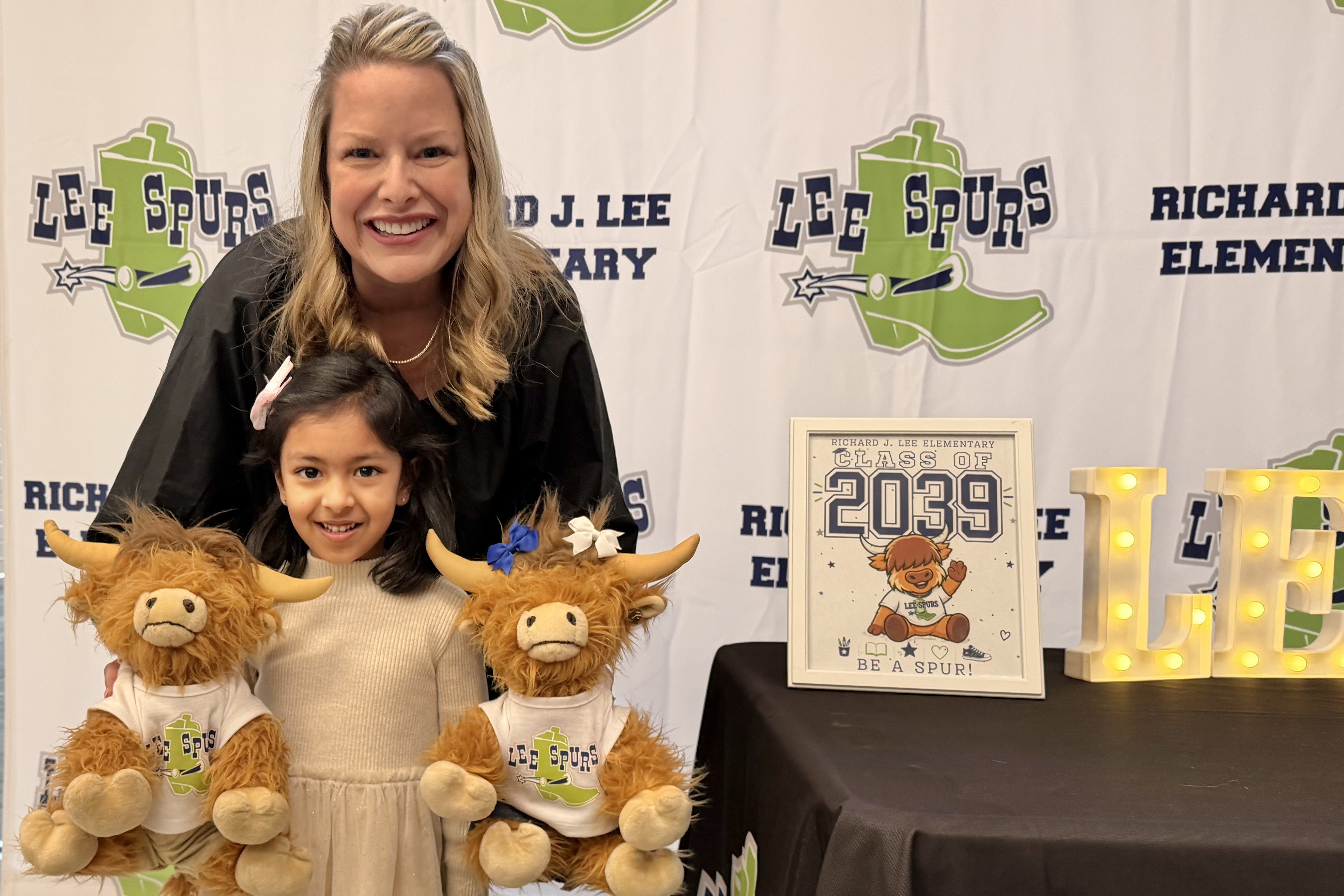 Lee Elementary principal smiles with a future kindergartener during Kinder Round Up, posing with school mascot stuffed animals in front of a Lee Spurs backdrop and Class of 2039 sign.