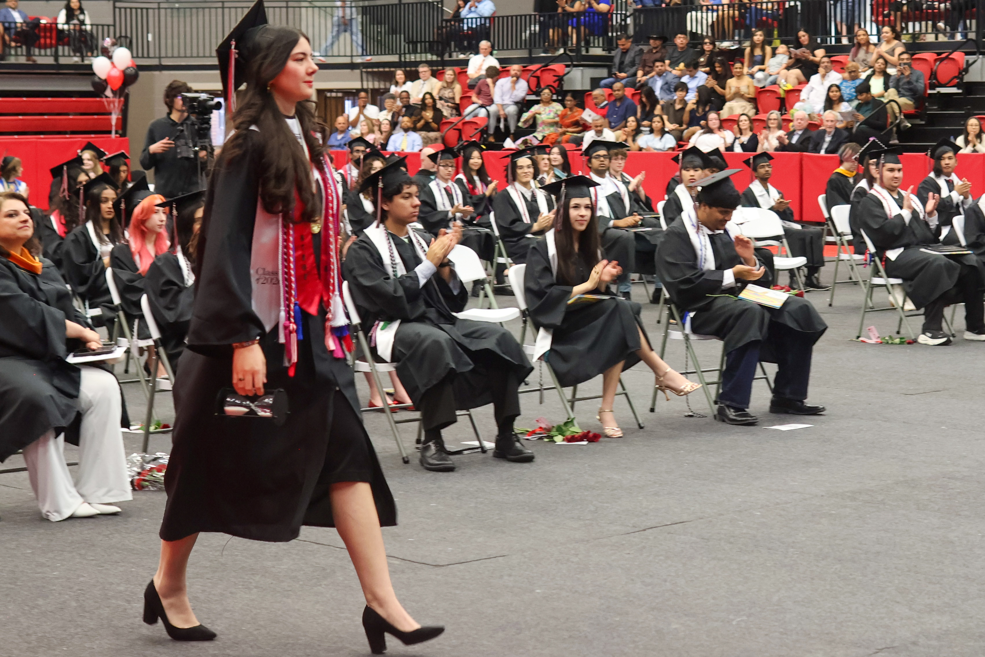 New Tech graduate in cap and gown walks across the ceremony floor as fellow graduates and guests applaud.