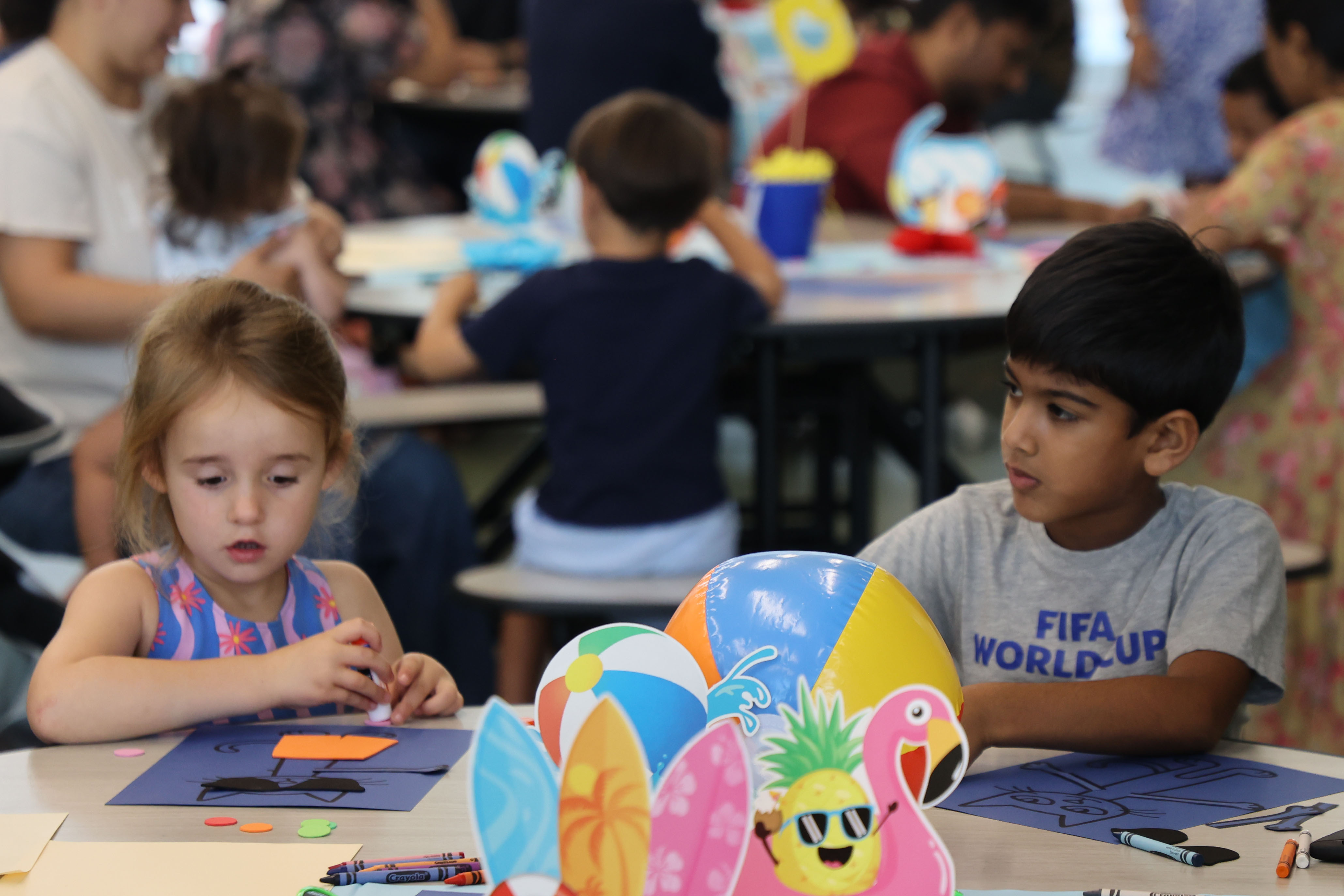 Two elementary-aged students work on a craft activity at a table decorated with colorful beach ball and tropical cutout centerpieces during a summer learning event, with other children and families engaged in similar activities in the background.