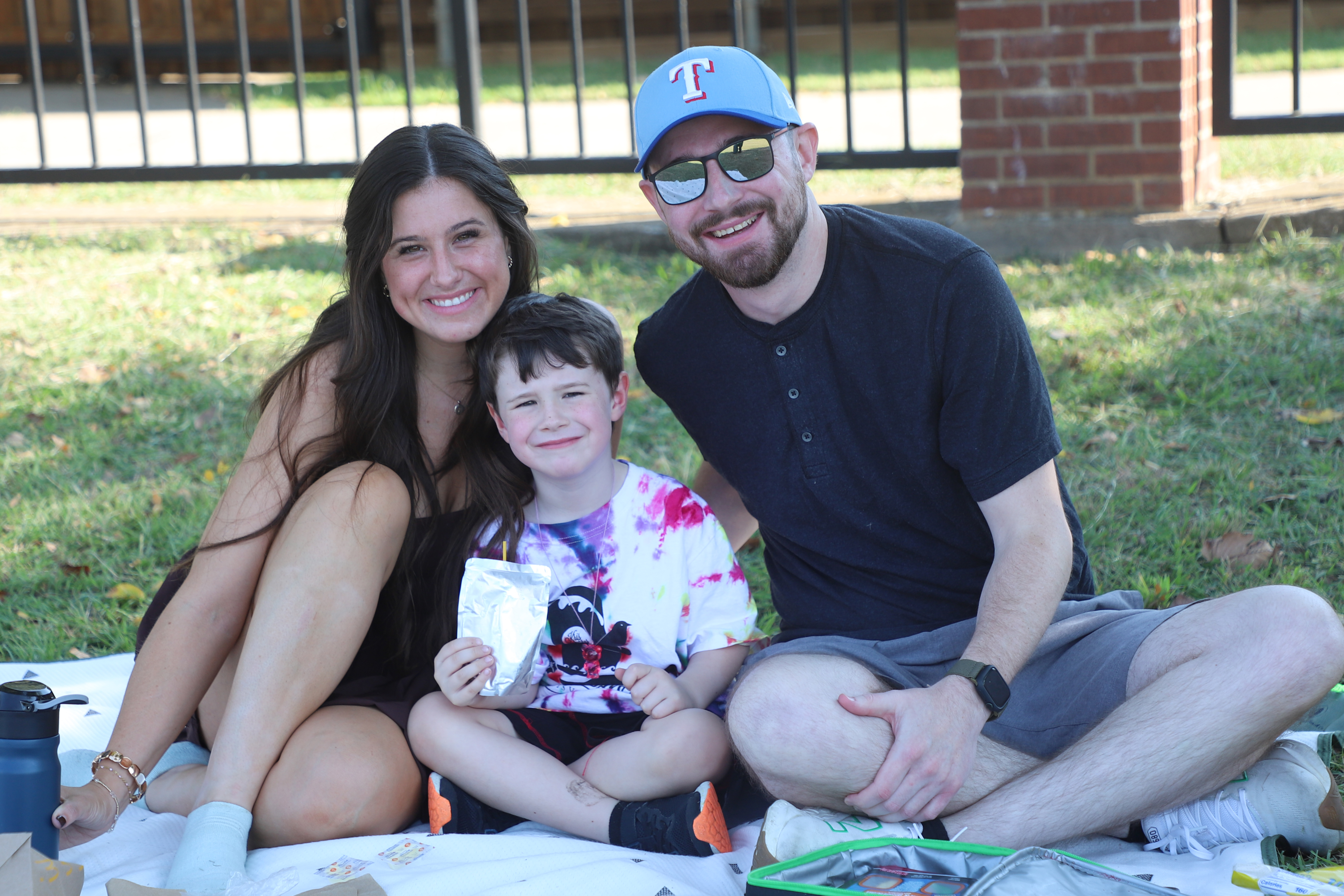 Smiling family of three sitting together on a picnic blanket outdoors, with a young child holding a drink pouch.