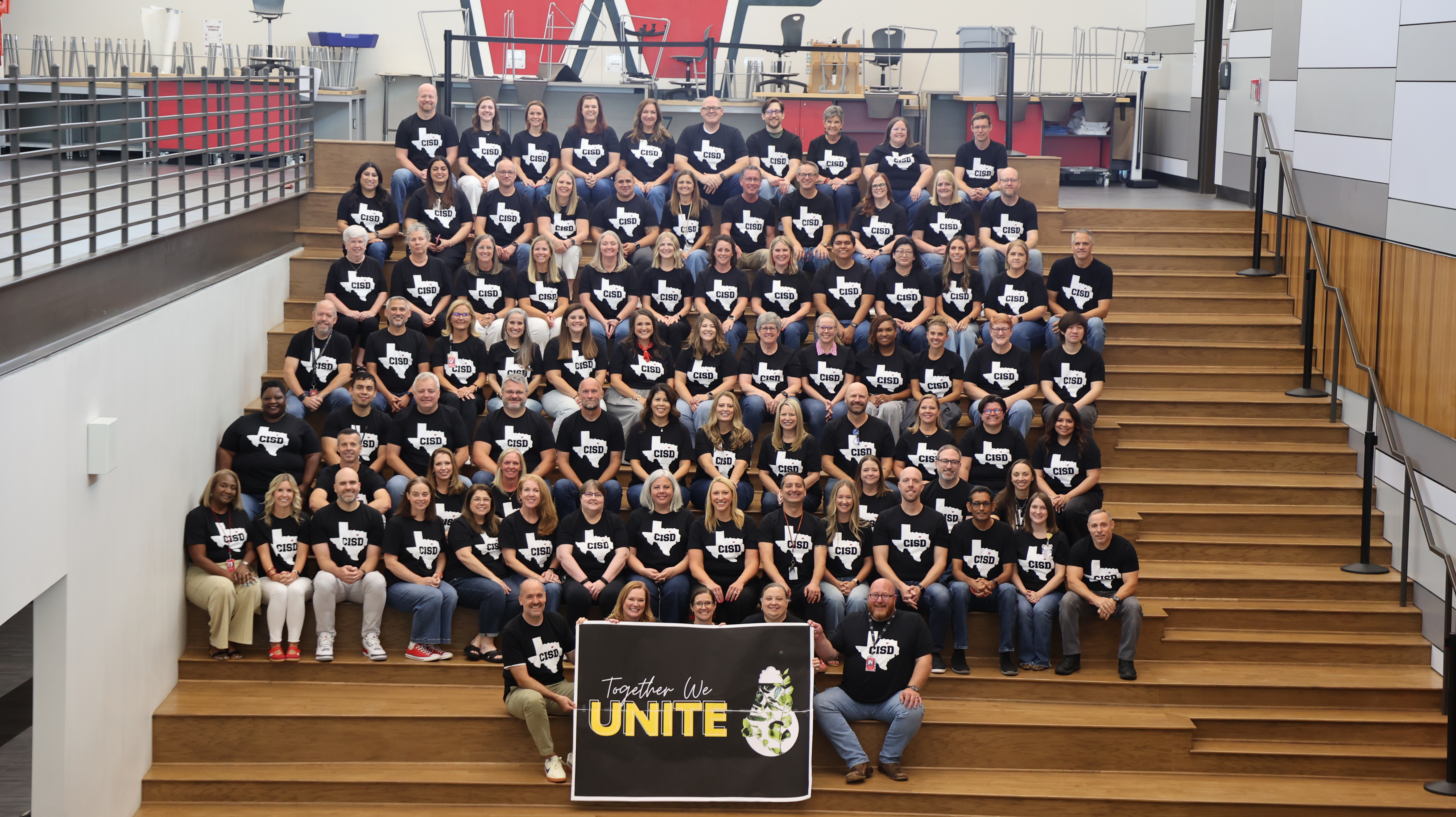 Group photo of a large staff (approximately 90 people), wearing matching black "CISD" T-shirts, seated on tiered wooden bleacher steps in a school building. Two people in front hold a sign reading "Together We Unite."