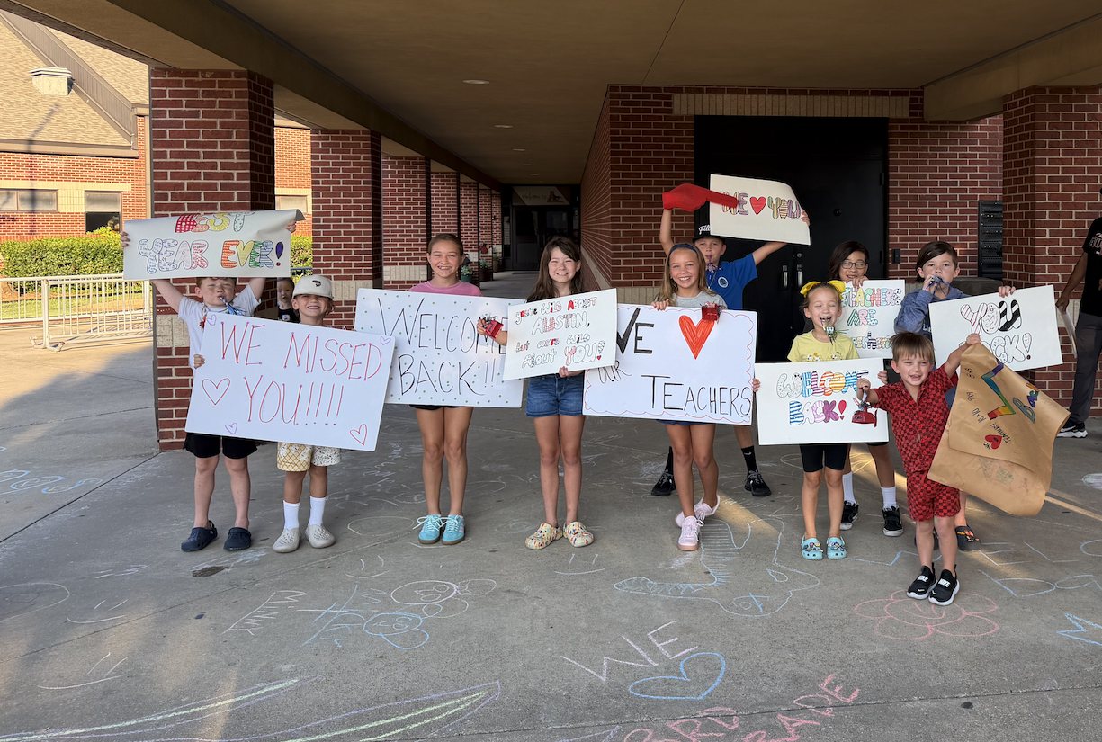 A group of smiling children hold handmade "Welcome Back" signs for teachers outside a school entrance decorated with sidewalk chalk art.