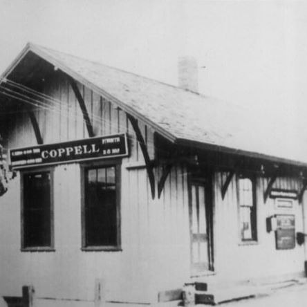 Historic black-and-white photo of the Coppell train depot, a wood-plank building with a peaked roof and a sign reading "Coppell."