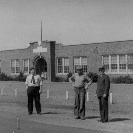 Vintage black-and-white photo of three men in hats standing in front of a brick school building with a flagpole.