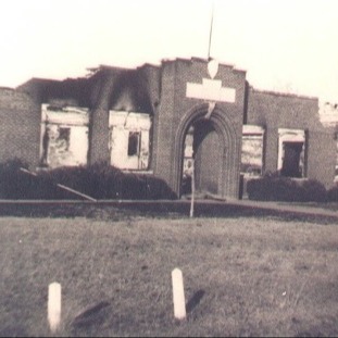 Historic photo of a fire-damaged brick school building with charred walls and a collapsed roof.