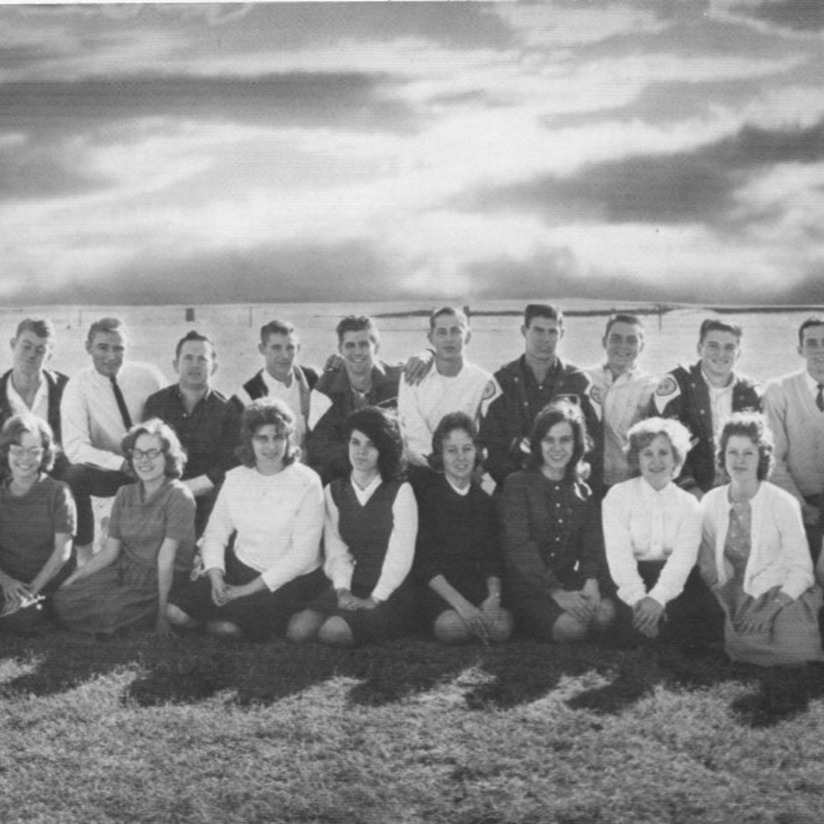 Black-and-white group photo of a first graduating class, young men standing and young women kneeling in two rows on a grassy field under a cloudy sky.