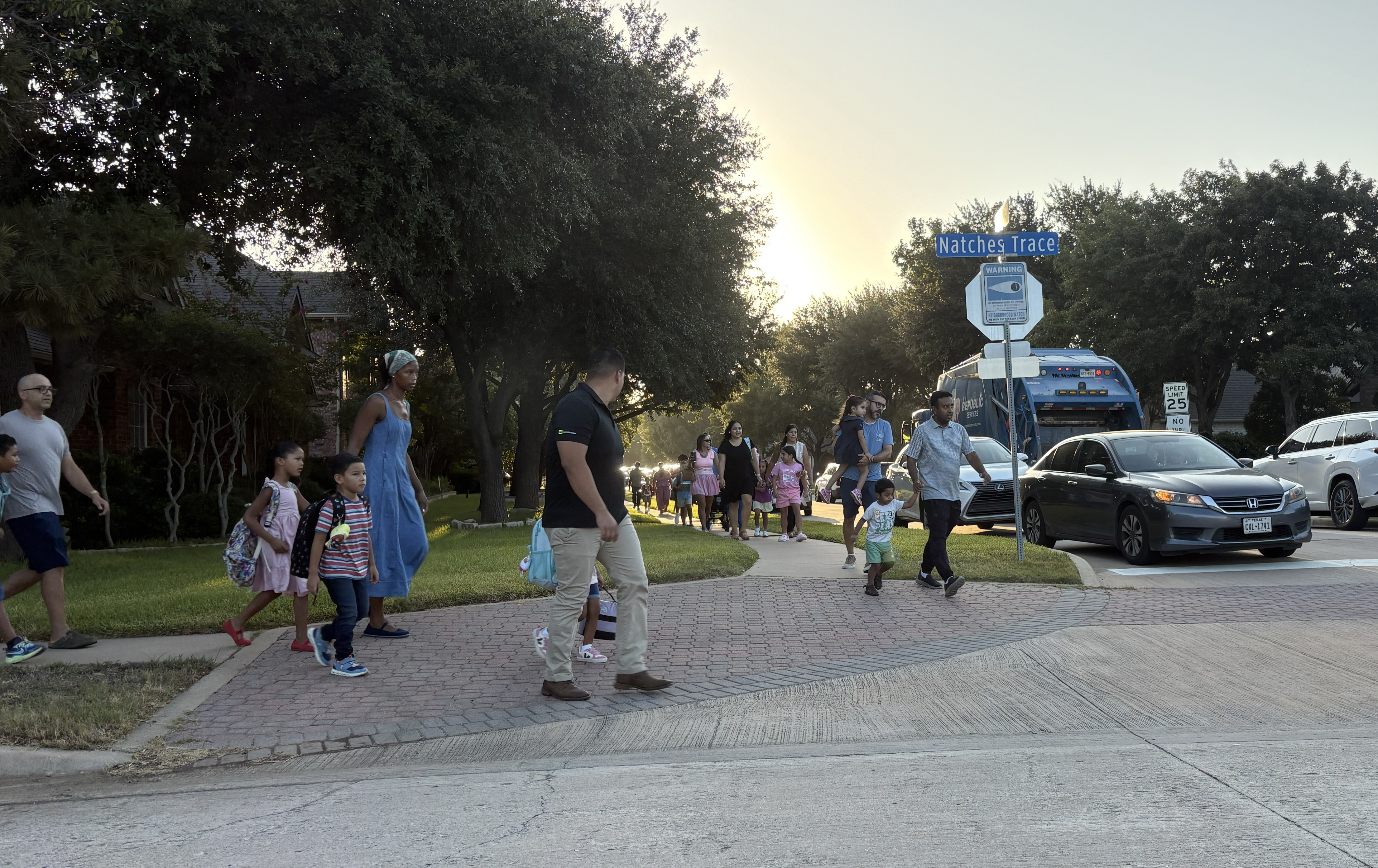 Families walk along a neighborhood sidewalk near a "Natches Trace" street sign at sunrise, with parents and children carrying backpacks heading toward school, cars parked along the curb.