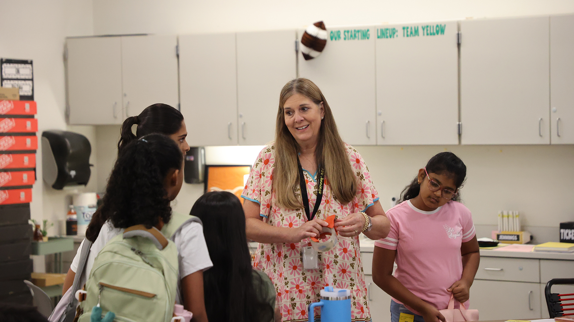 A woman in a floral dress talks with students gathered around her in a classroom.