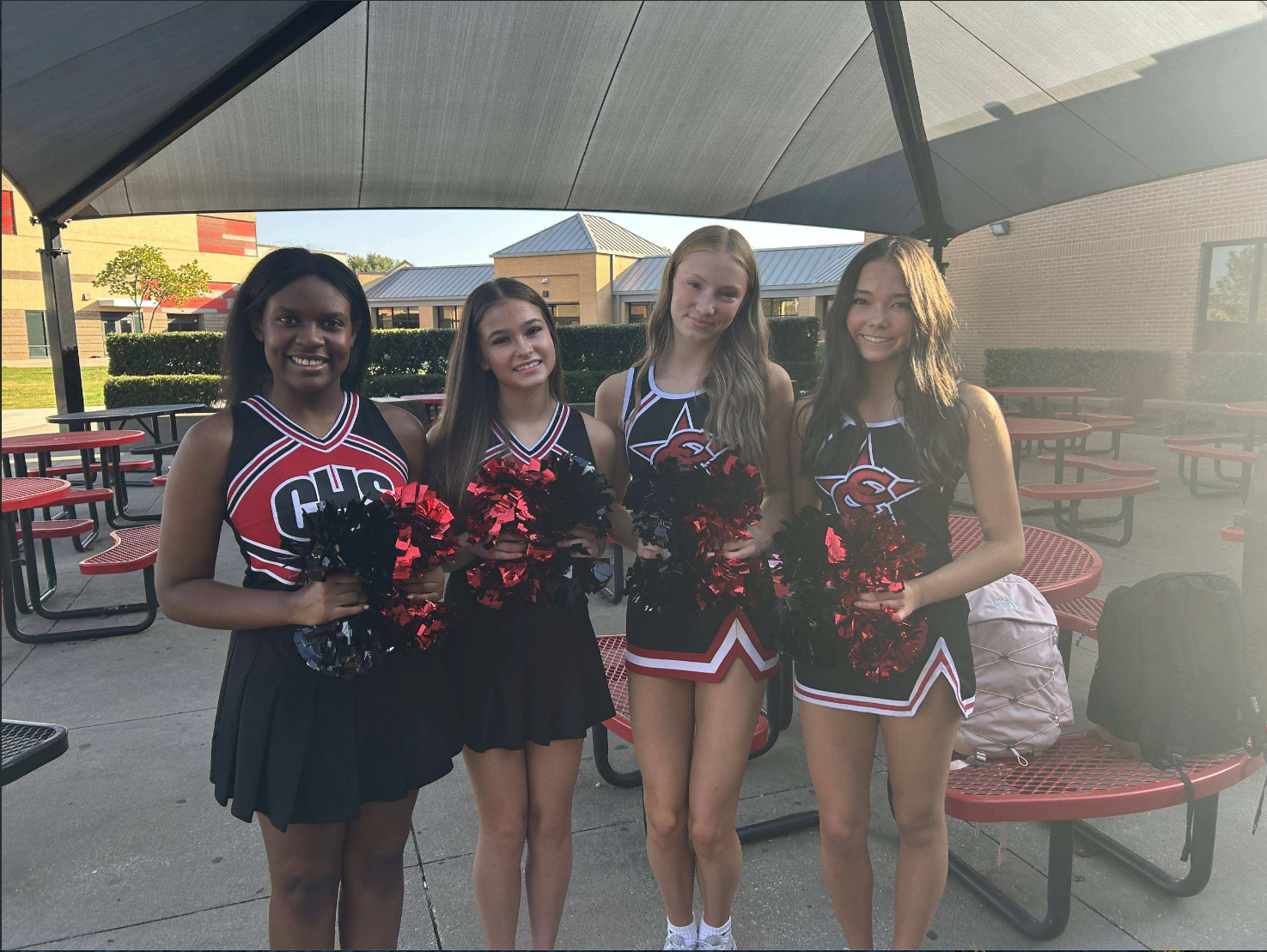 Four cheerleaders in matching red, black, and white uniforms pose with pom-poms outdoors at a school courtyard.