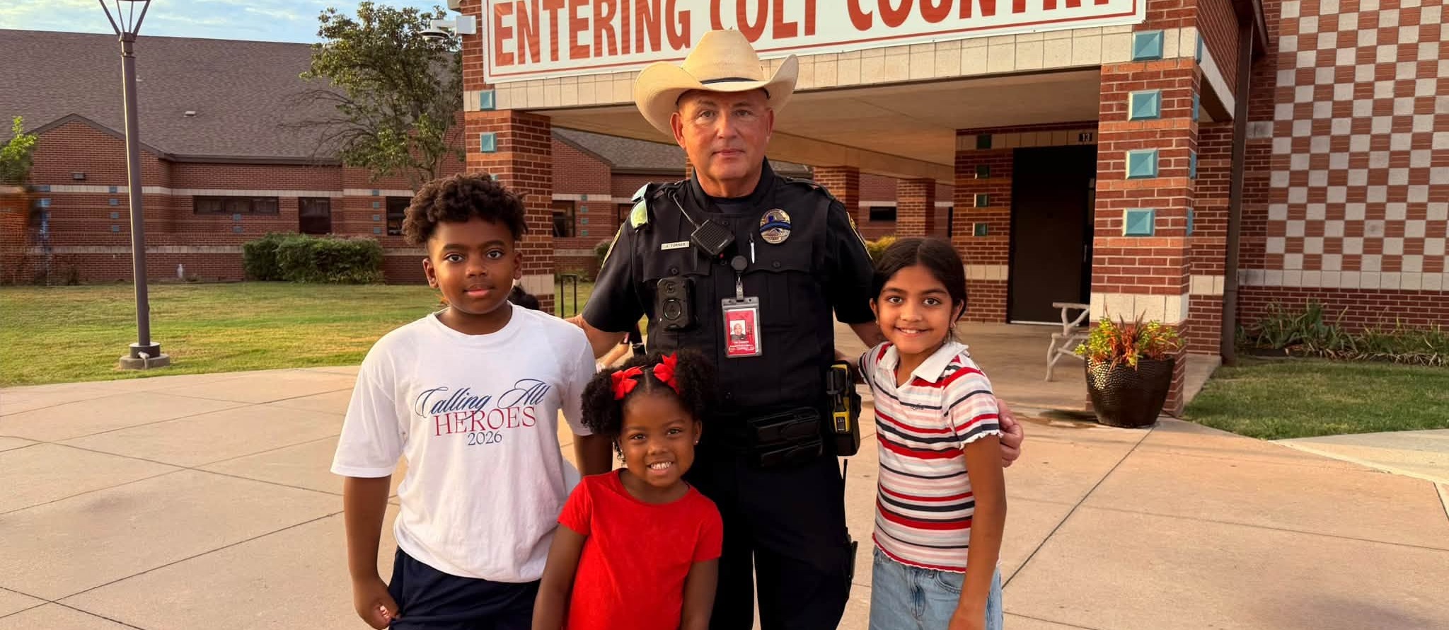 Three children pose with a police officer outside a brick school building with a “Entering Colt Country” sign.