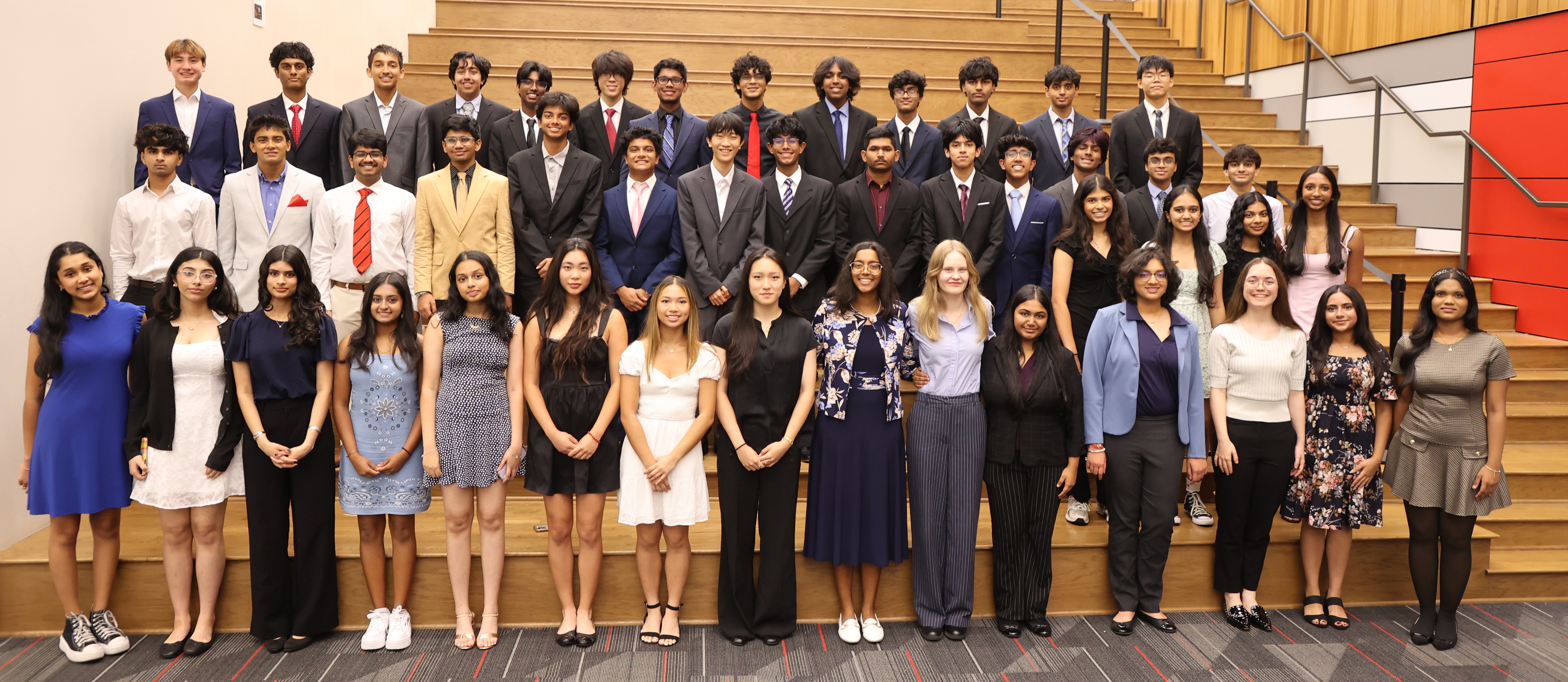 Group photo of National Merit Semifinalists from Coppell High School and New Tech, posed in three rows on a wooden amphitheater-style staircase beneath the red and white Coppell sign. About 45 students stand together in formal attire, with the boys in suits and ties in the back and middle rows, and the girls in dresses and blouses in the front row.