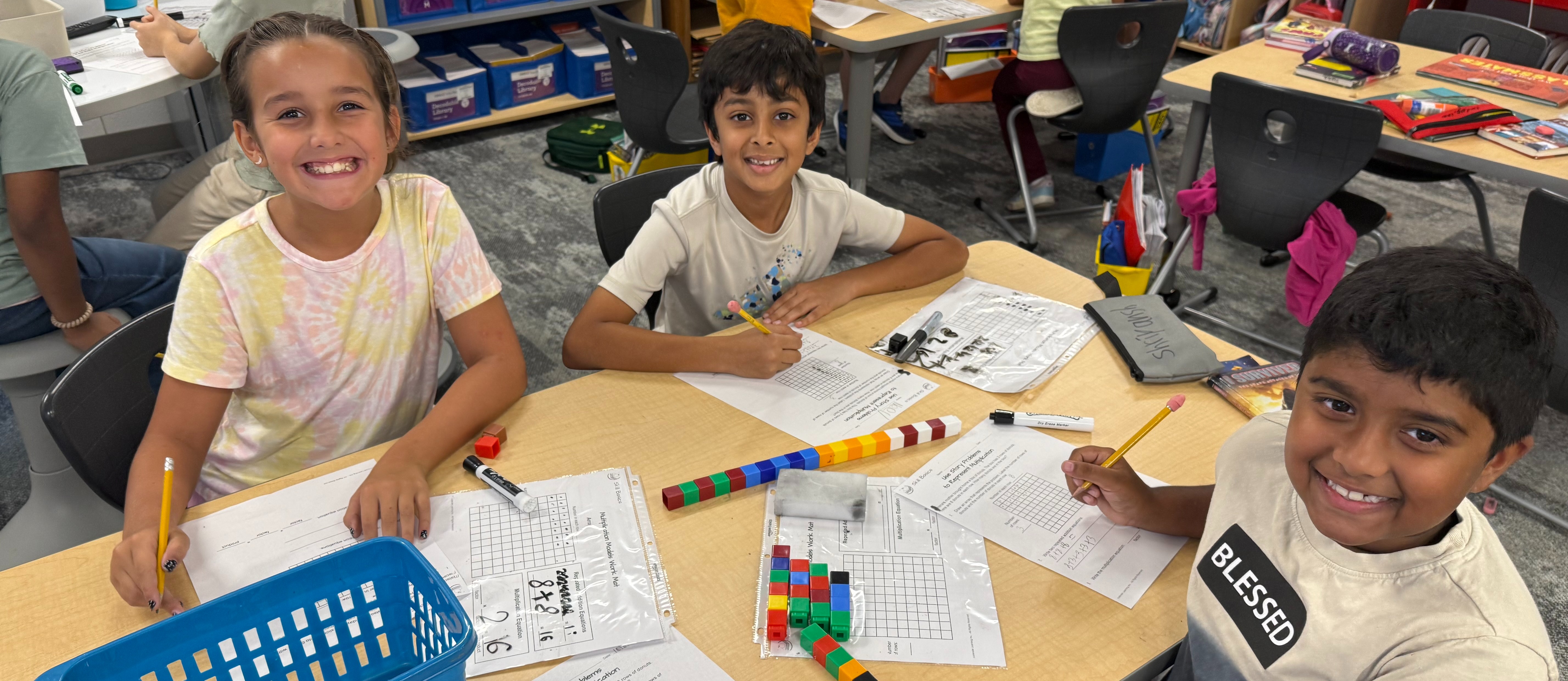 Three smiling third-grade-aged students sit together at a classroom table, looking up at the camera while holding pencils. In front of them are multiplication array worksheets in plastic sleeves, a blue basket, dry-erase markers, and colorful linking cubes stacked in rows and a long bar. Behind them, other students work at tables near shelves of labeled book bins.