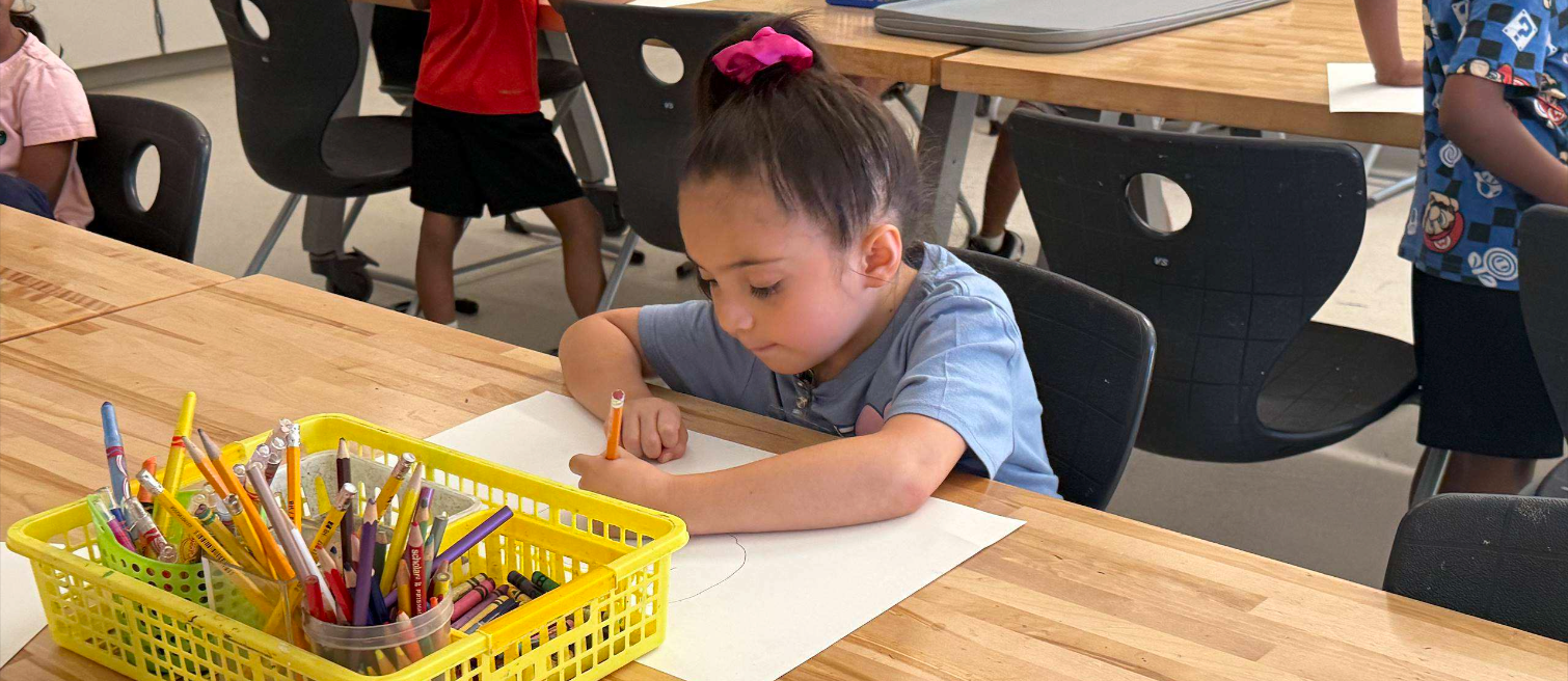 A young girl with dark hair pulled up in a pink scrunchie sits at a wooden classroom table, leaning over a sheet of white paper as she draws with an orange pencil. A yellow basket of pencils, crayons, and markers sits beside her. Other students work at tables in the background.