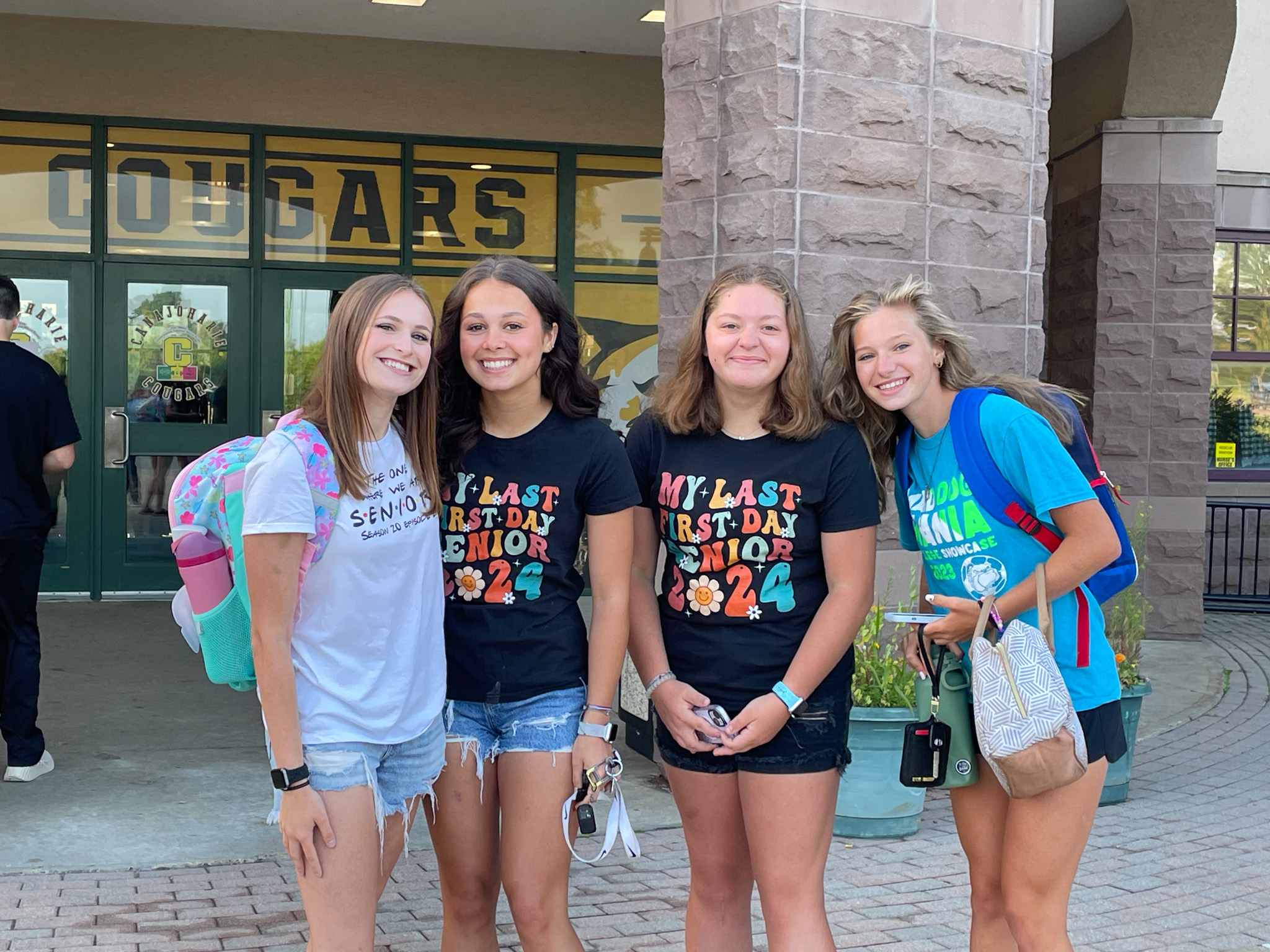 Four students, each wearing Last First Day and Senior 2024 shirts, pose in front of the high school entrance. 