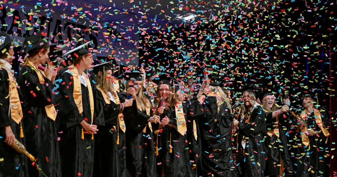 Graduates in black robes with yellow sashed celebrate as confetti rains down. 