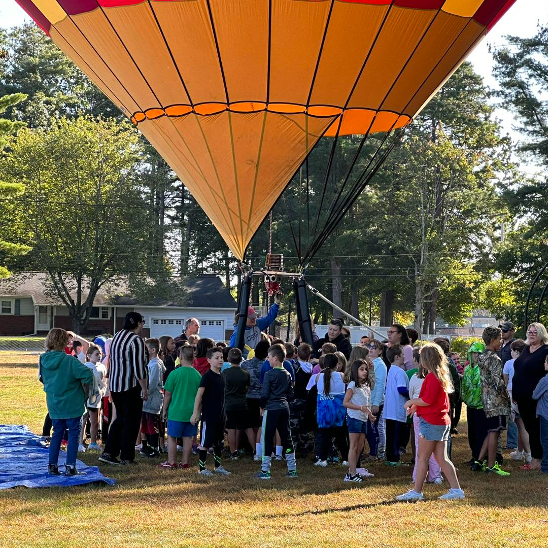 A hot air balloon sits in the field by the school. Students observe it. 