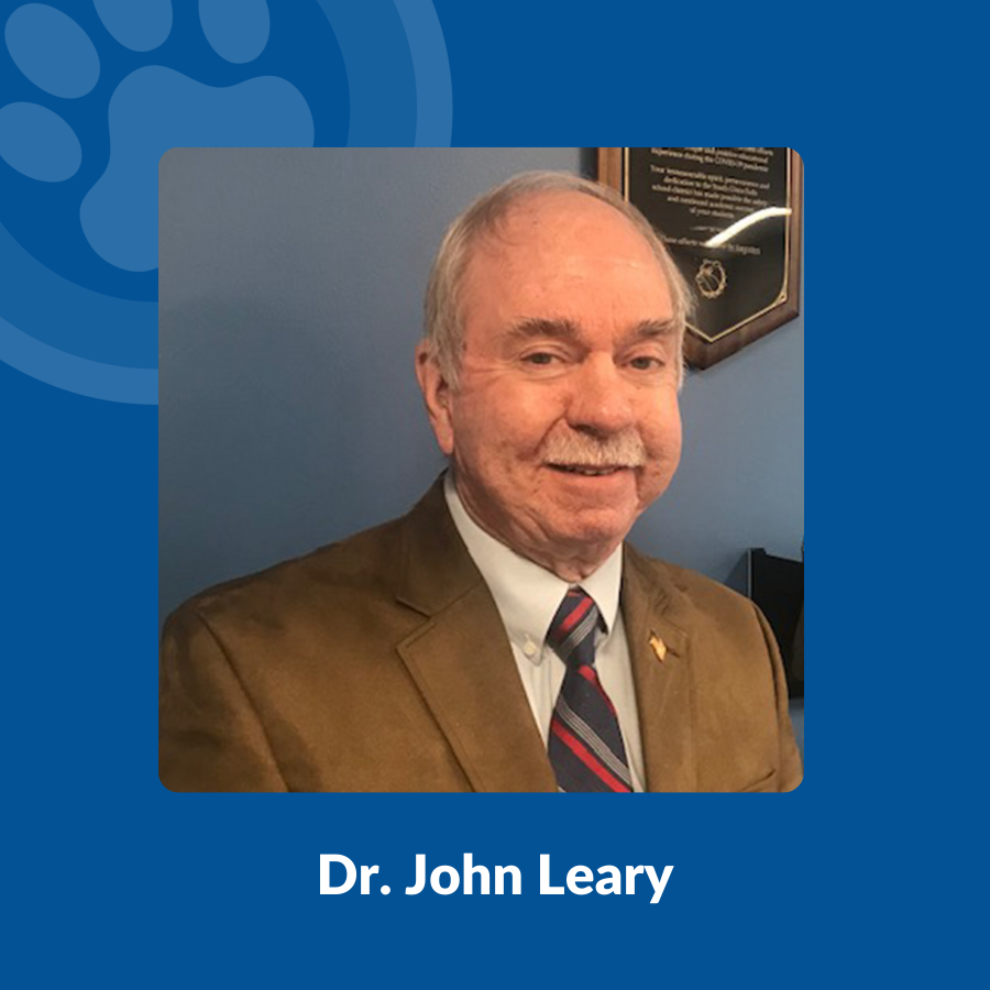 Dr. John Leary wears a brown suit while standing against a blue wall