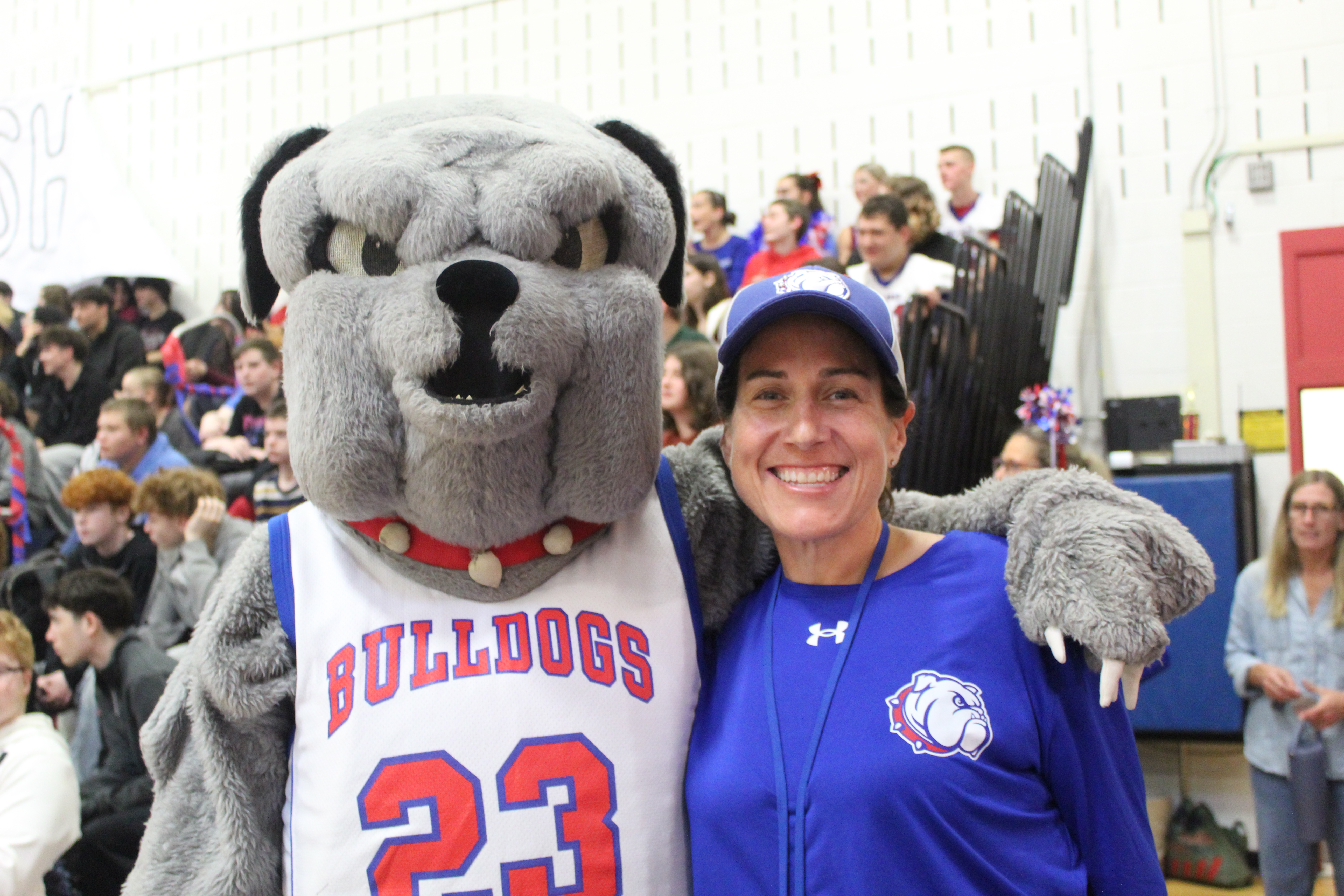 A smiling woman wearing a blue shirt and baseball cap poses with a large grey bulldog mascot in a gymnasium.