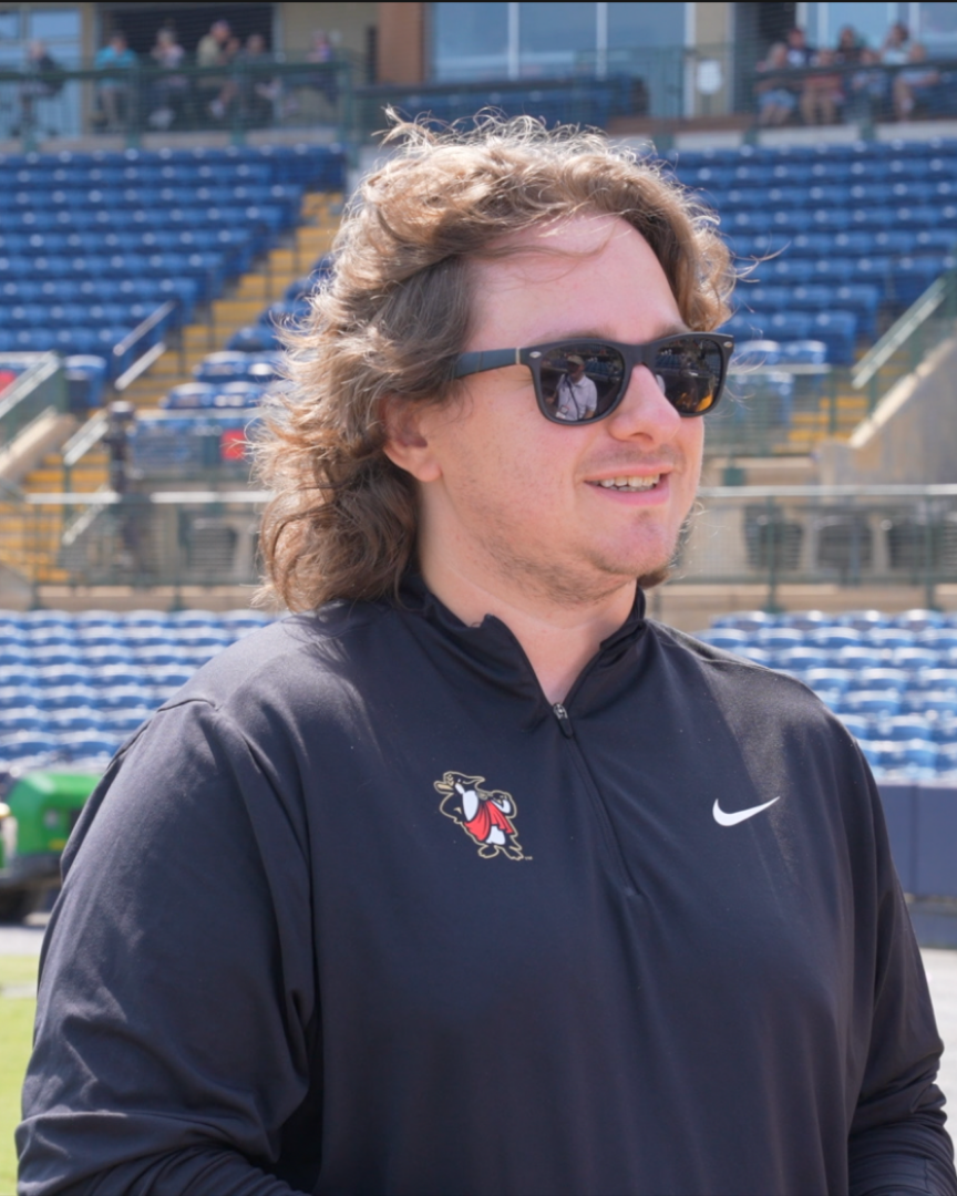 Man with long hair, sunglasses, and black shirt stands in a stadium with empty blue seats.