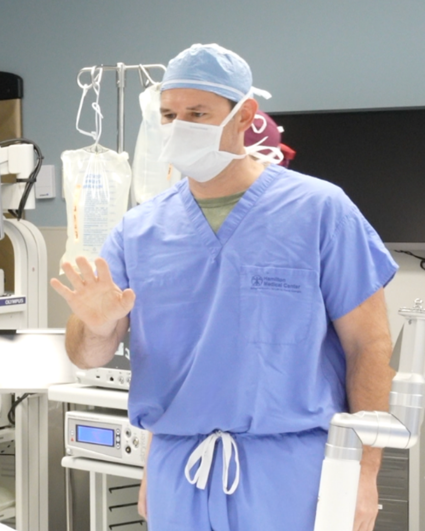 A medical team in blue scrubs and masks in a hospital room. One person wears a VR headset.