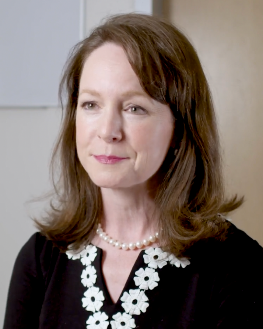 A woman in a black top with white floral trims and a pearl necklace sits in front of a white wall.