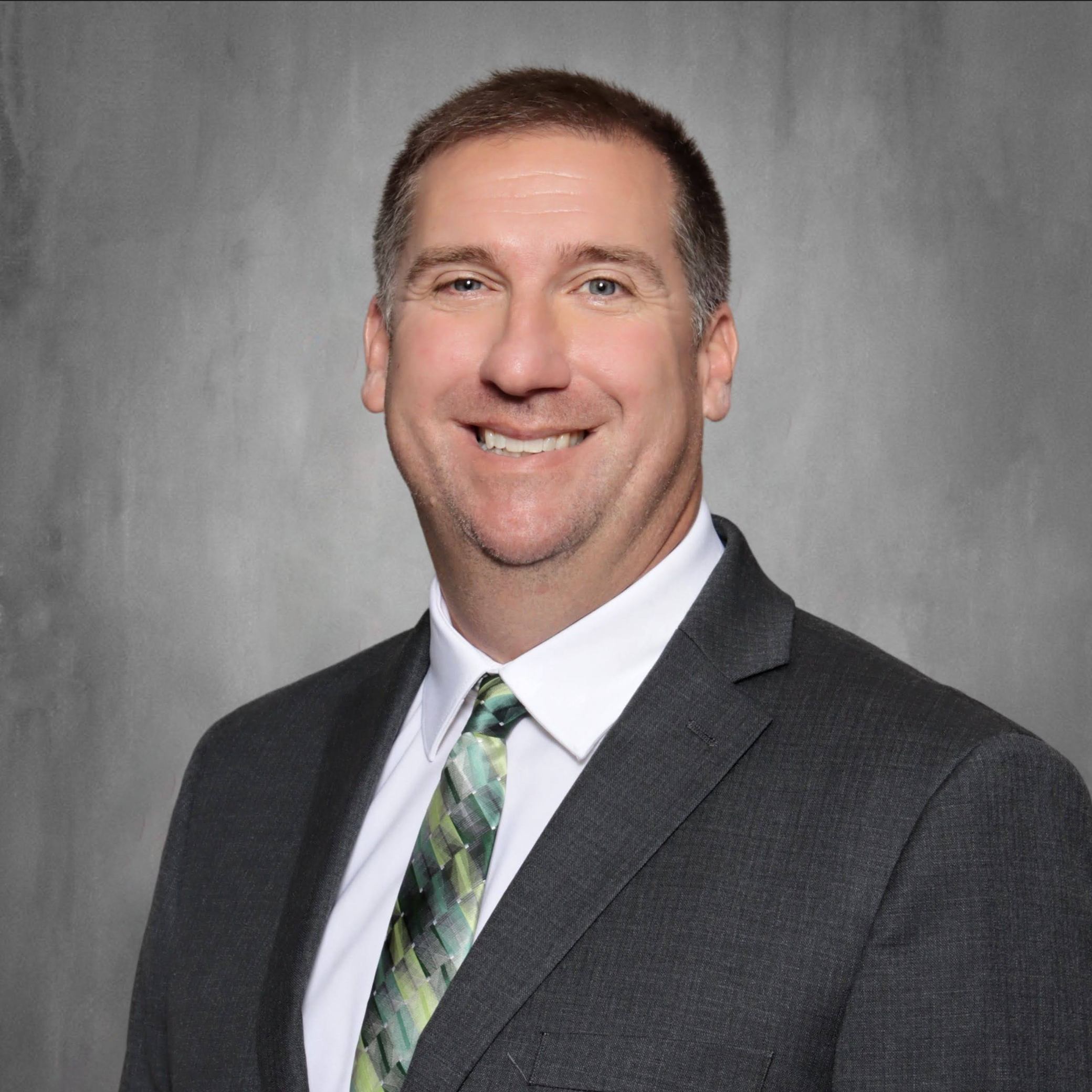 Man with short dark hair, wearing a light gray suit and a light green tie, standing against a light gray background, smiling.