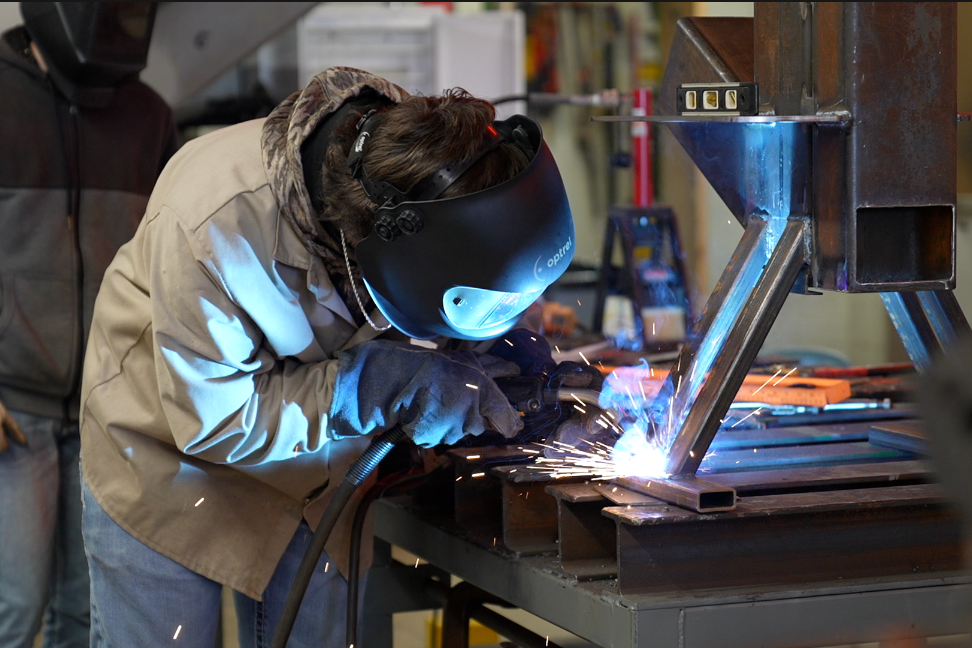 A person in protective gear welding on a metal structure in a workshop. Another person stands nearby.