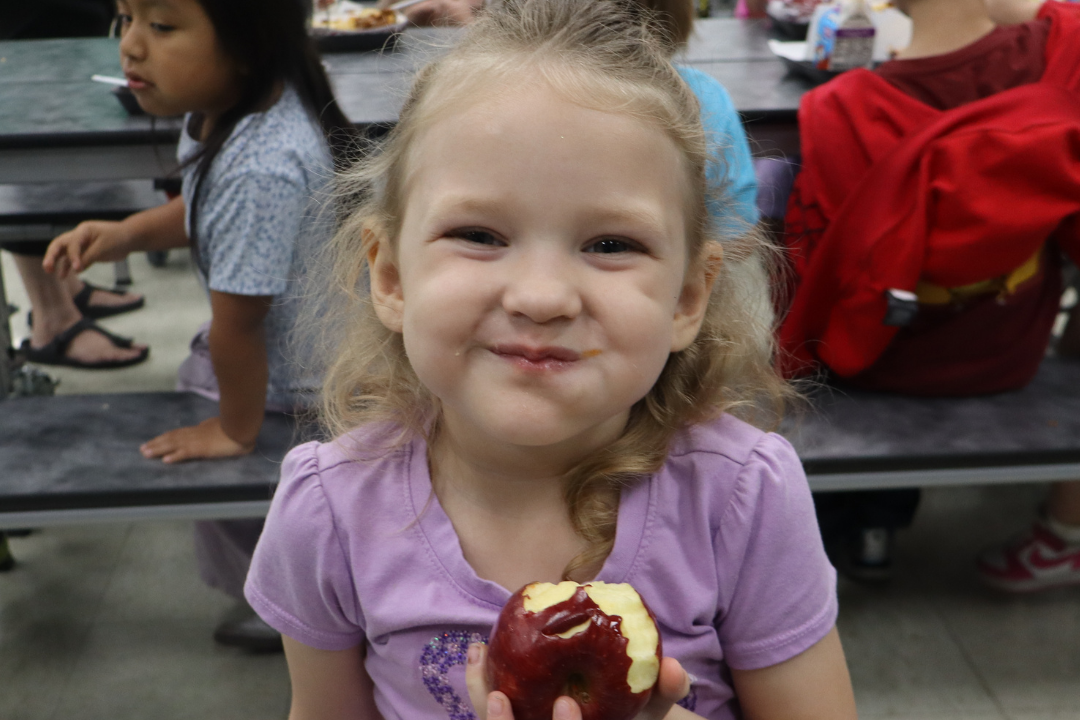 A girl with blonde hair sits at a table, holding a bitten apple. Behind her, other children sit at tables.