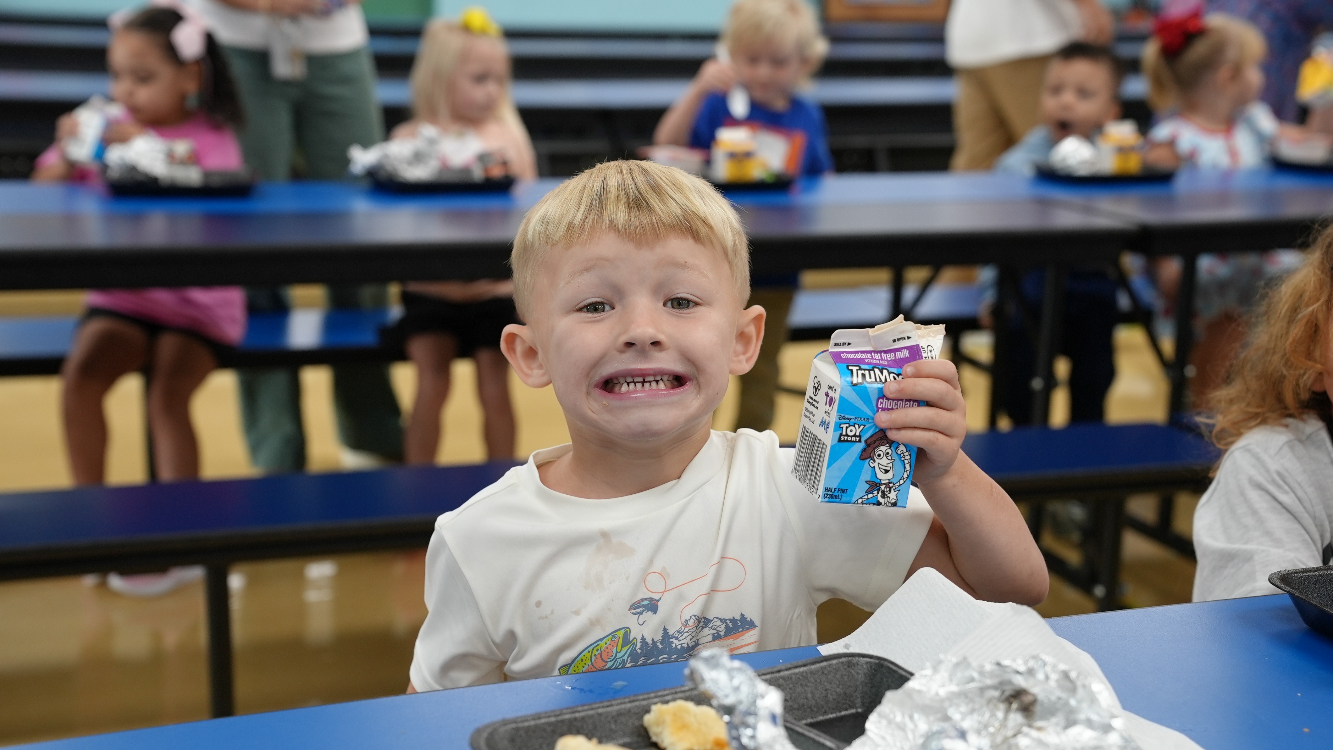 Blonde boy seated at a blue table with lunch tray holding a carton of milk.