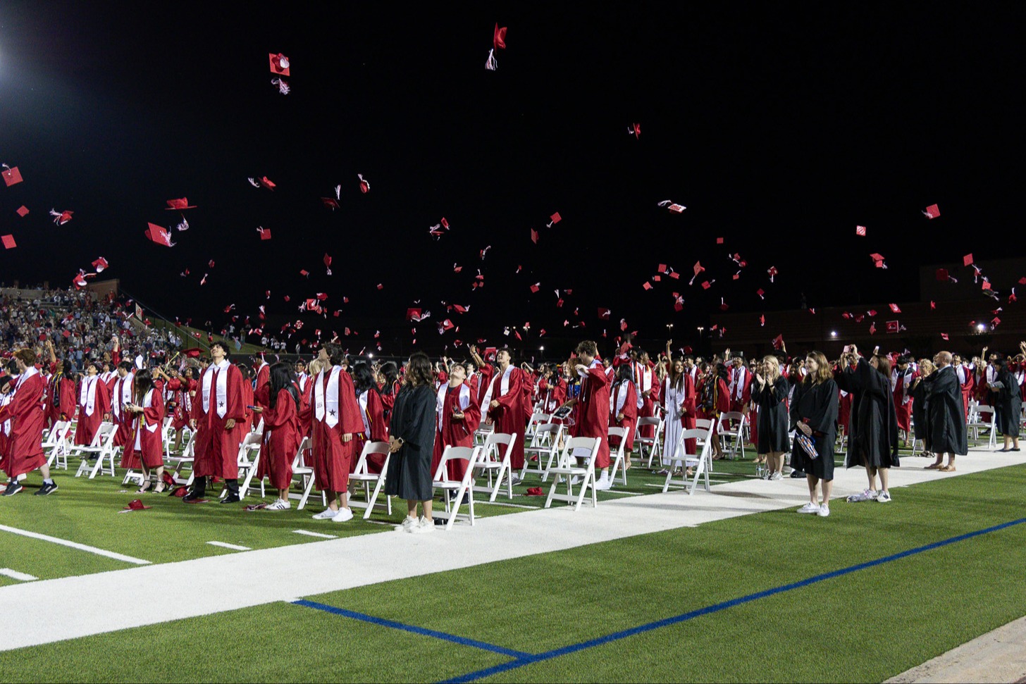 Graduates in red caps and gowns toss their caps into the night sky during an outdoor stadium graduation ceremony.